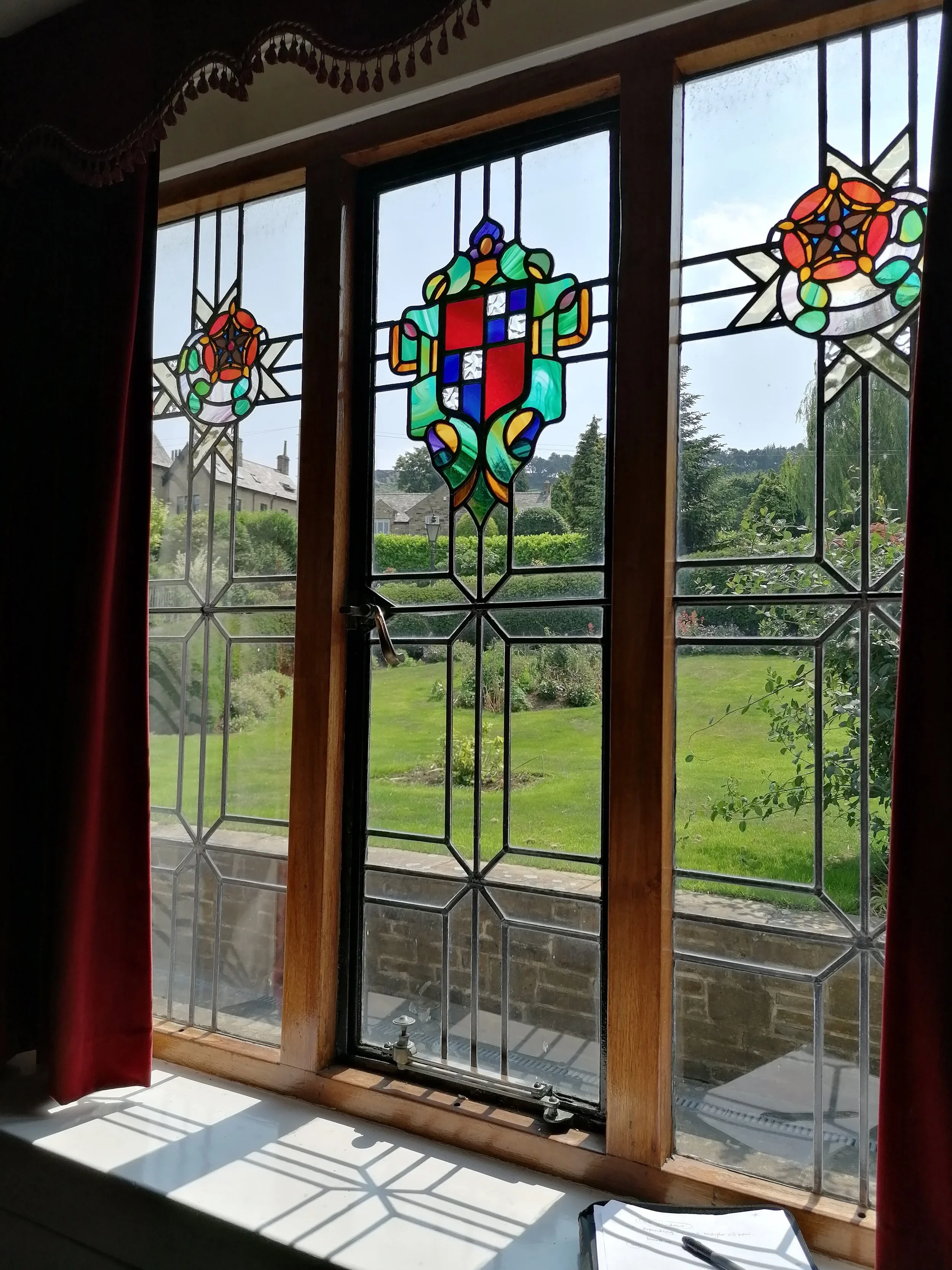 Interior view of a window with stained glass featuring a colorful shield and floral patterns, outside a garden with greenery and houses.