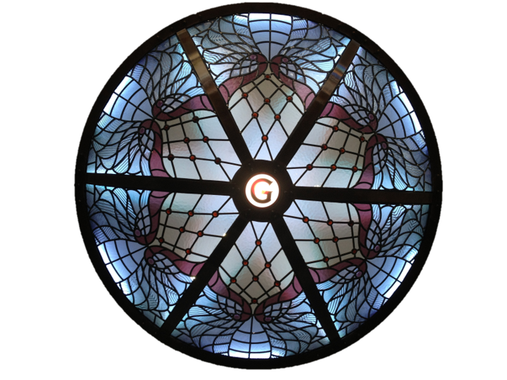 A circular stained glass window featuring intricate blue, pink, and white designs with a central letter G and segmented sections.