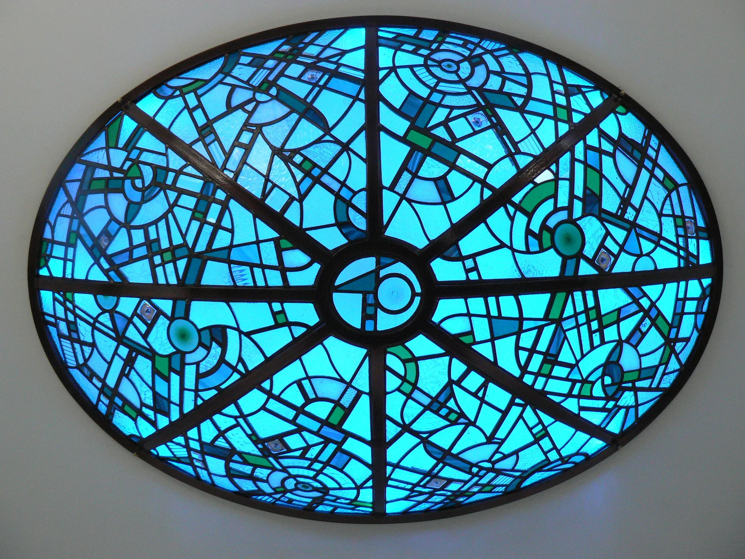 A stained glass window with an abstract, geometric pattern in shades of blue and green, shaped as a semi-circle.