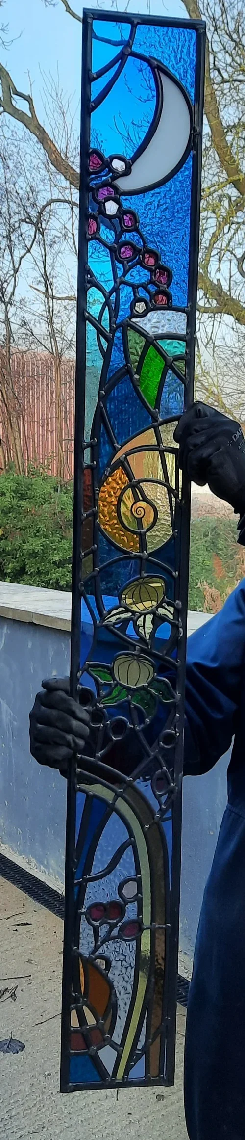A stained glass panel depicting a crescent moon, purple flowers, green leaves, a spiral pattern, and abstract shapes in blue, orange, and yellow, held outdoors against a backdrop of trees and a fence.