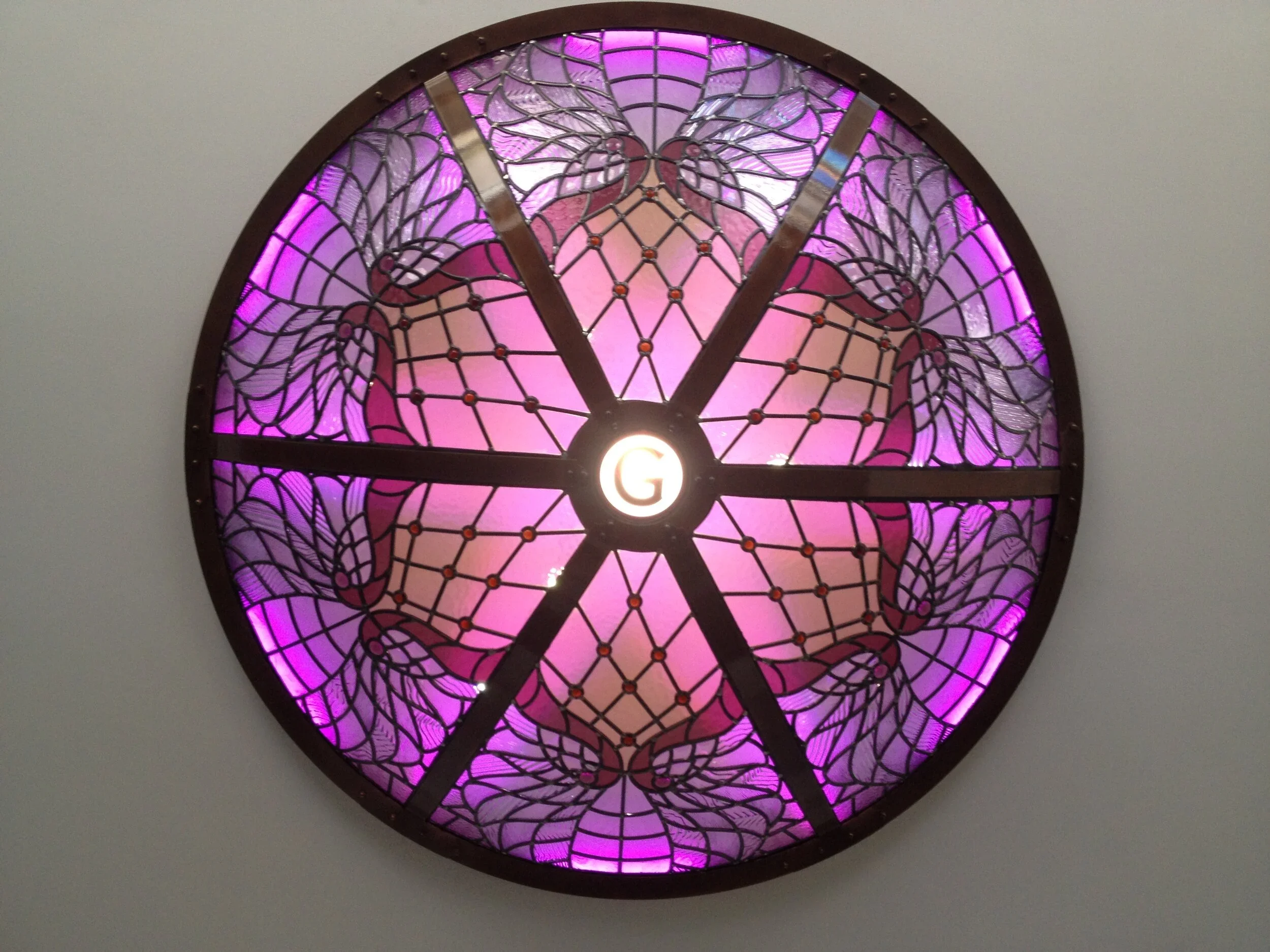 Stained glass window with a purple and pink butterfly pattern, surrounded by a metal frame, with the letter 'G' illuminated at the bottom center.