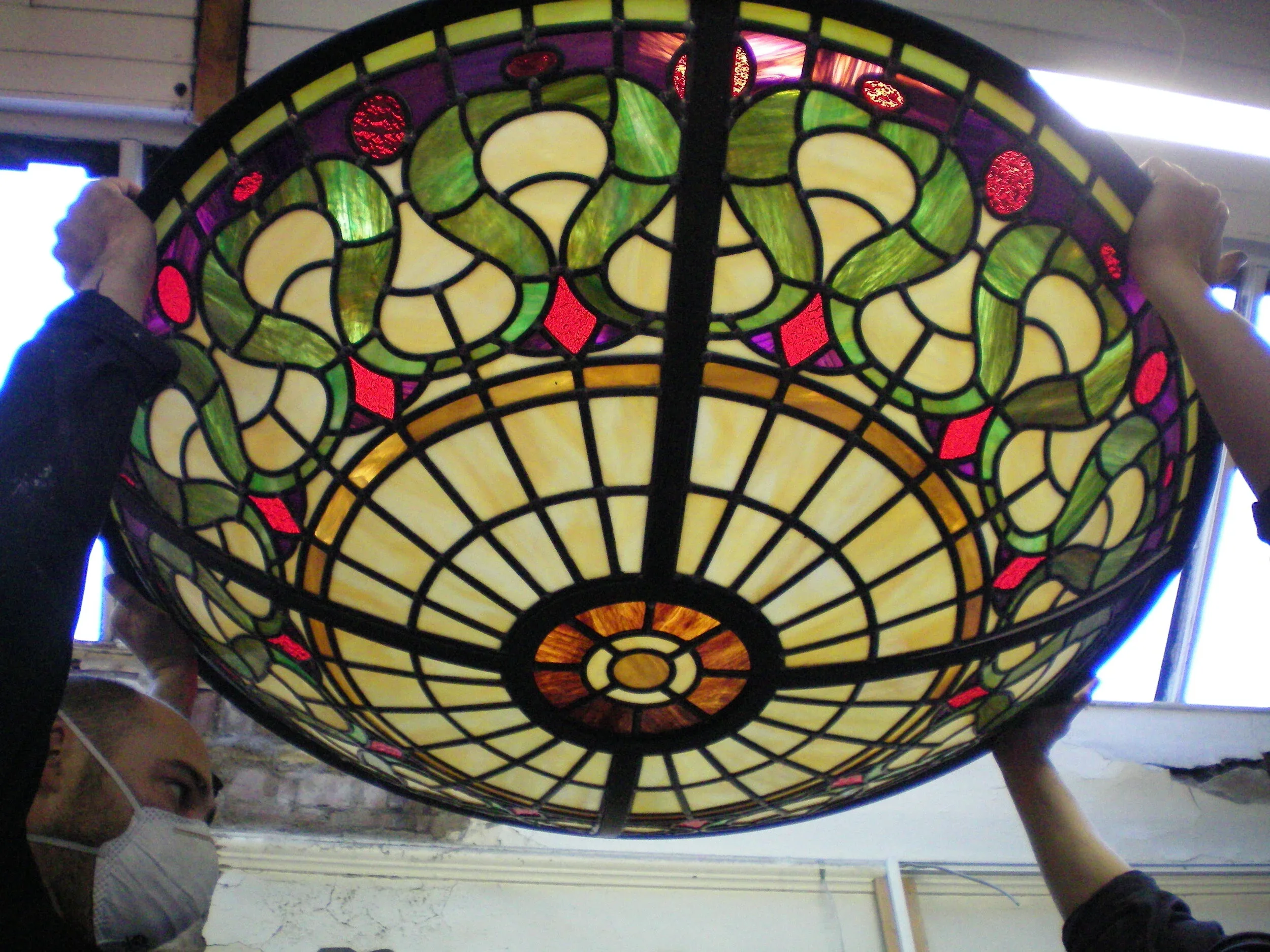 People holding a stained glass lamp with floral and geometric designs, featuring yellow, green, purple, and red colors.