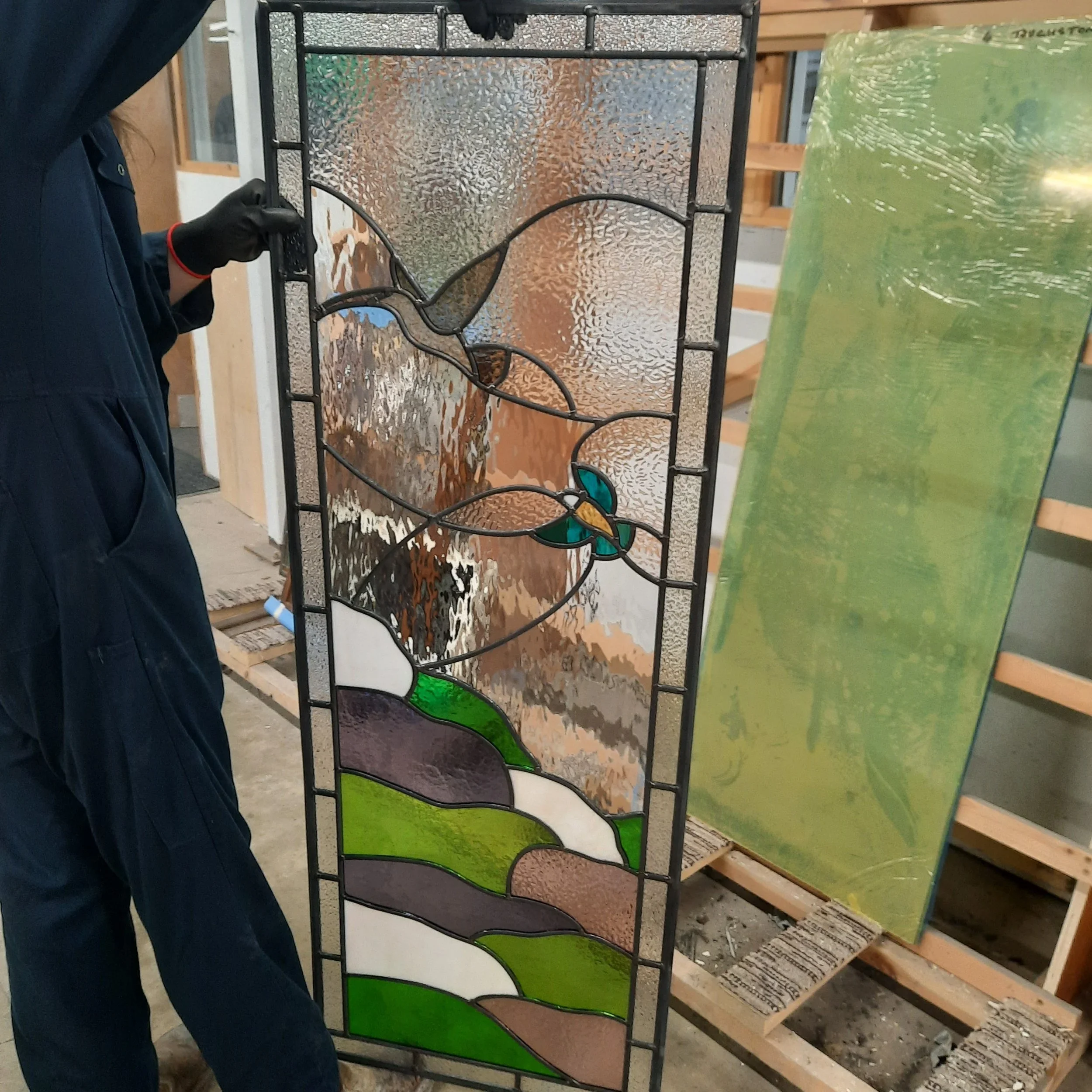 A person holding a stained glass panel featuring a bird design, with a green and brown landscape background.