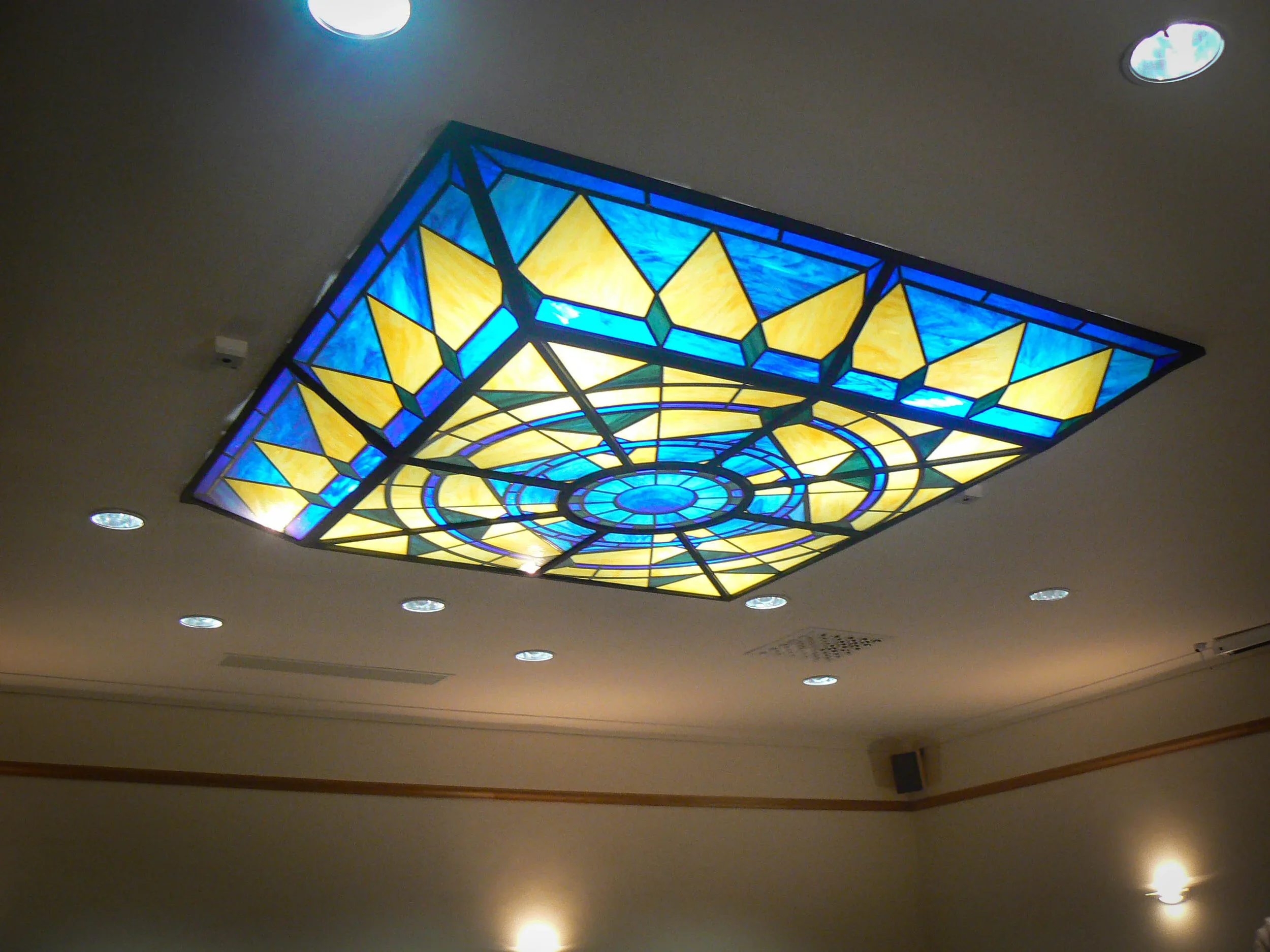 A colorful stained glass ceiling light fixture with geometric patterns in shades of blue, yellow, and green, mounted on a white ceiling with small recessed lights around it.