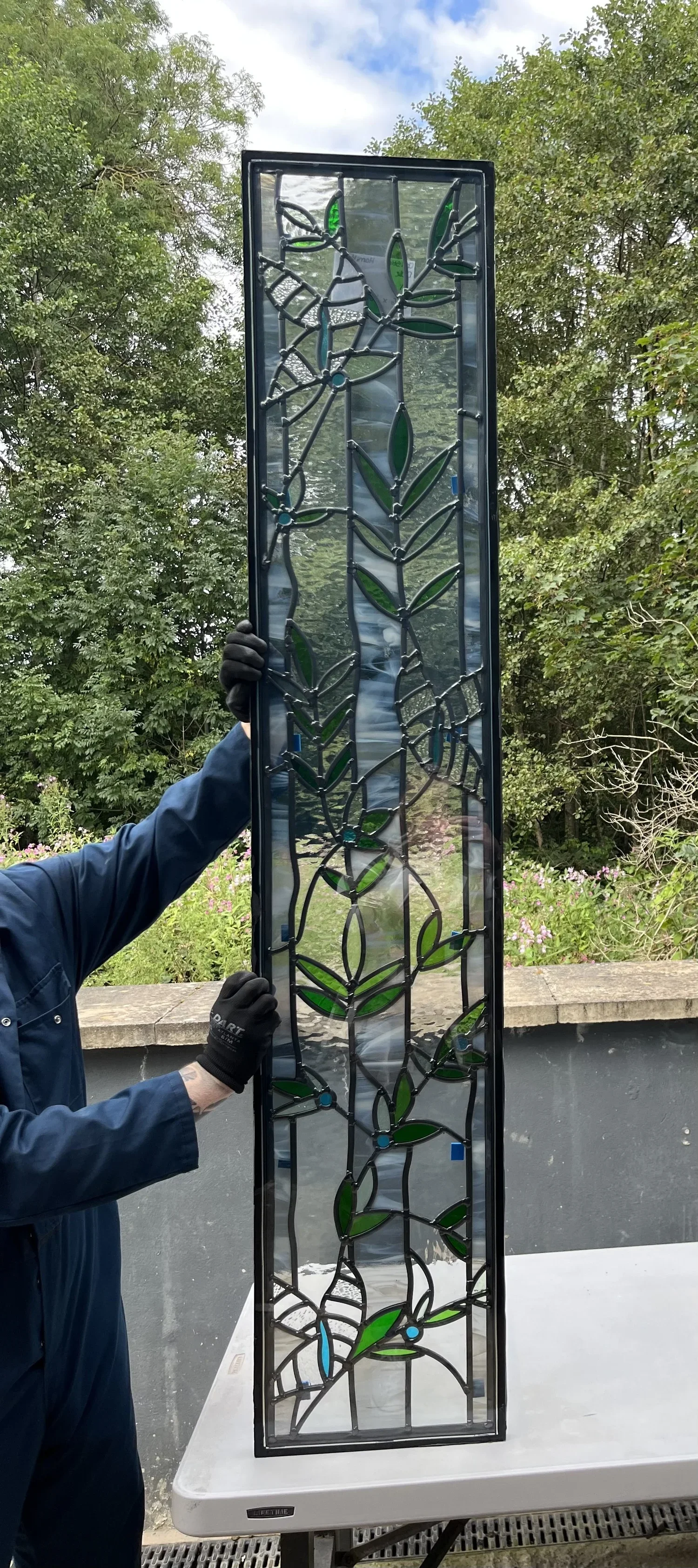 A person holding a tall stained glass panel with a floral design outdoors, with green trees and a cloudy sky in the background.