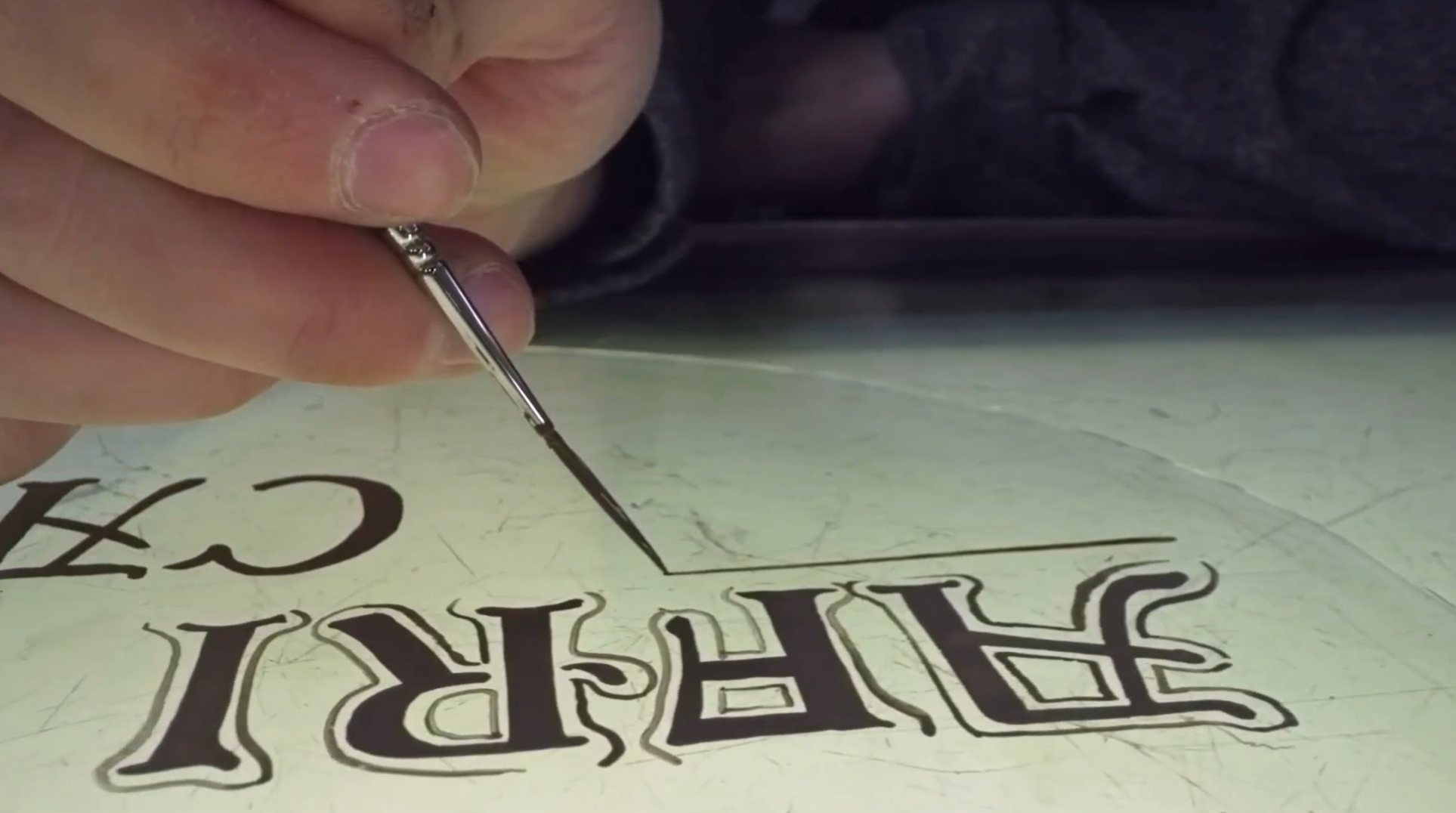 Close-up of a person using a fine brush to paint black outline details on a sign that reads "HAPPY ADVENTURE" in decorative letters.