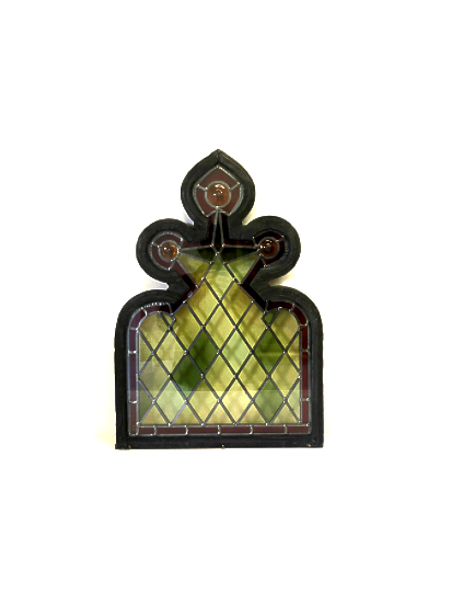 A stained glass window with a black frame, featuring a decorative top with three circular ornaments and a cross in the center. The window has a grid pattern with yellow and green glass panes, and offers a view of trees and sky outside.