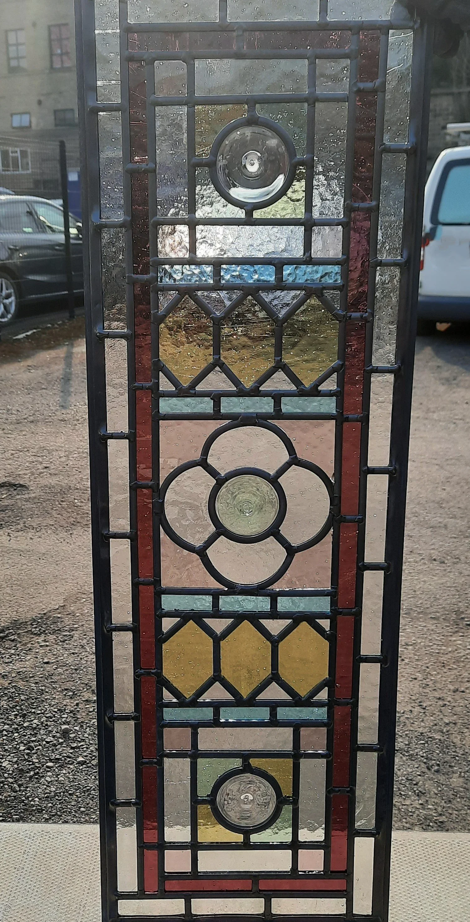 A stained glass decorative panel with geometric patterns and circular designs, featuring various colors including red, yellow, blue, and clear glass, set against an outdoor background with parked cars and a building.