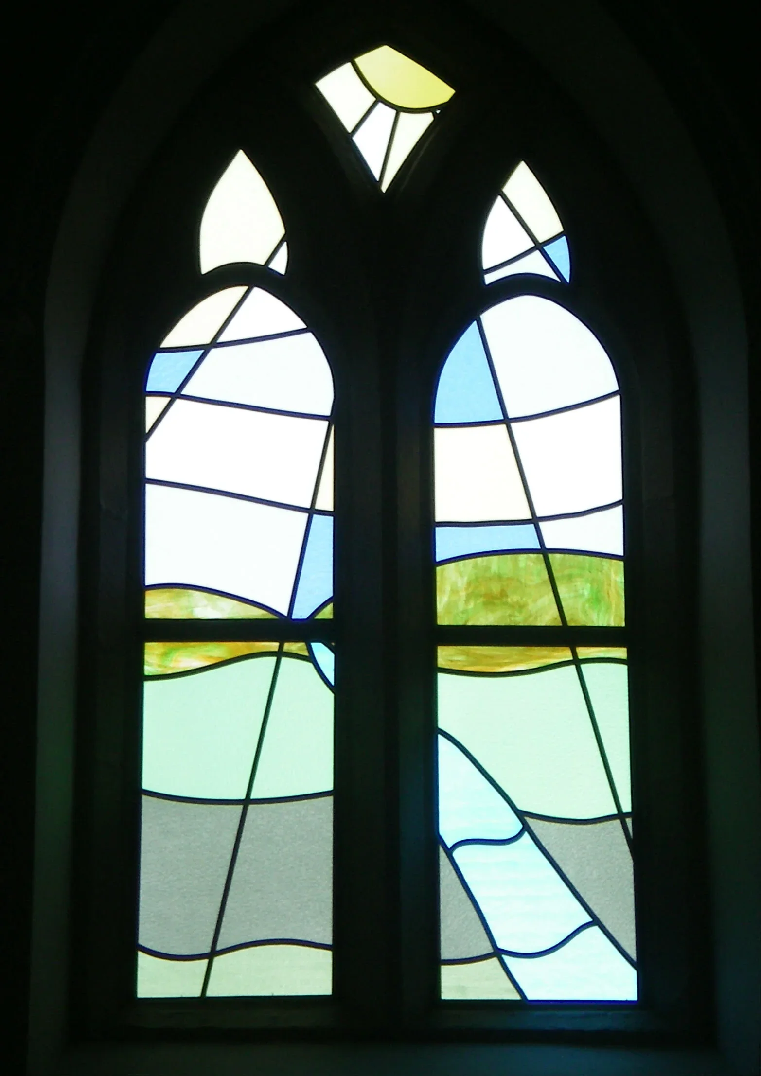 Colorful stained glass window with abstract geometric shapes in shades of blue, green, yellow, and white, framed in dark metal.