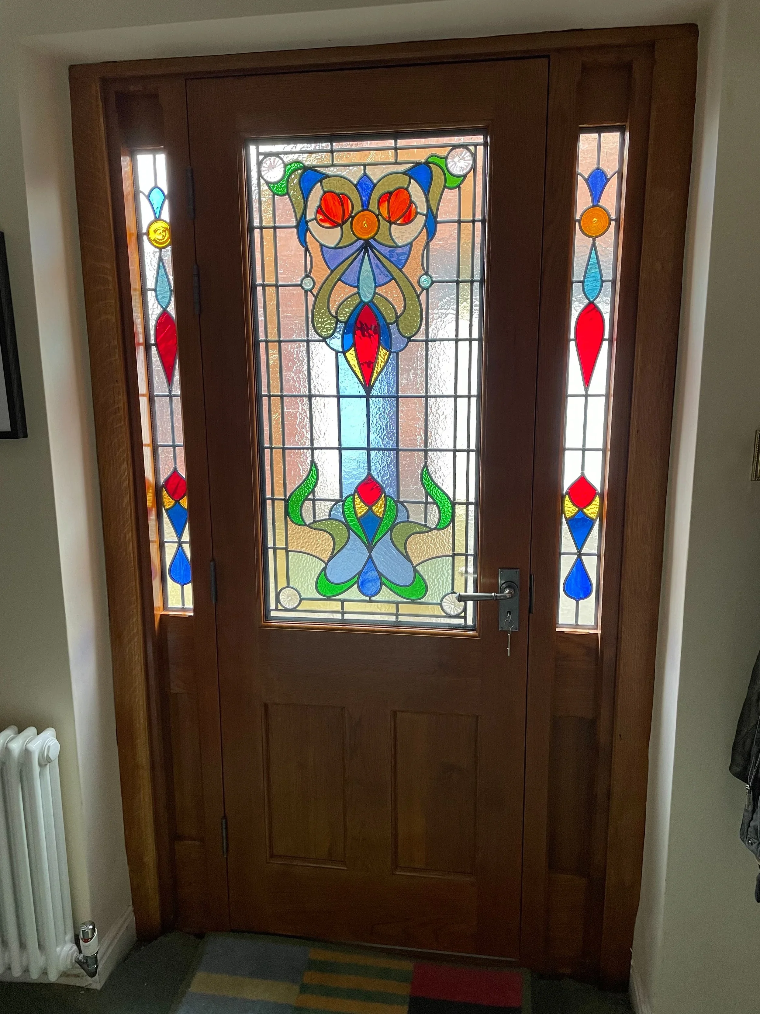 Wooden front door with stained glass window featuring a colorful floral and abstract design, next to a white radiator on the left side.