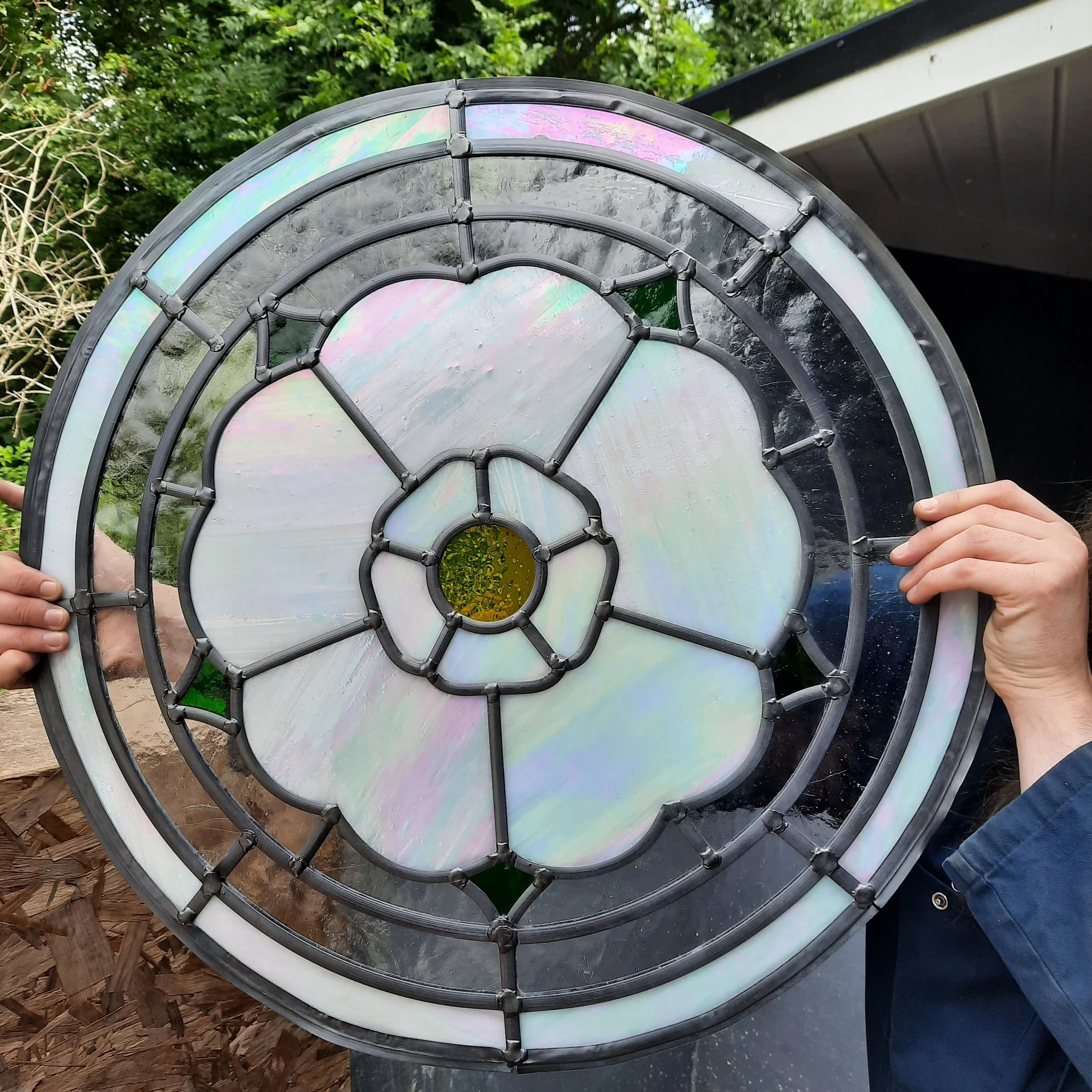 Close-up of a stained glass window with an iridescent or holographic effect held up by multiple people outside, with trees and a building in the background.