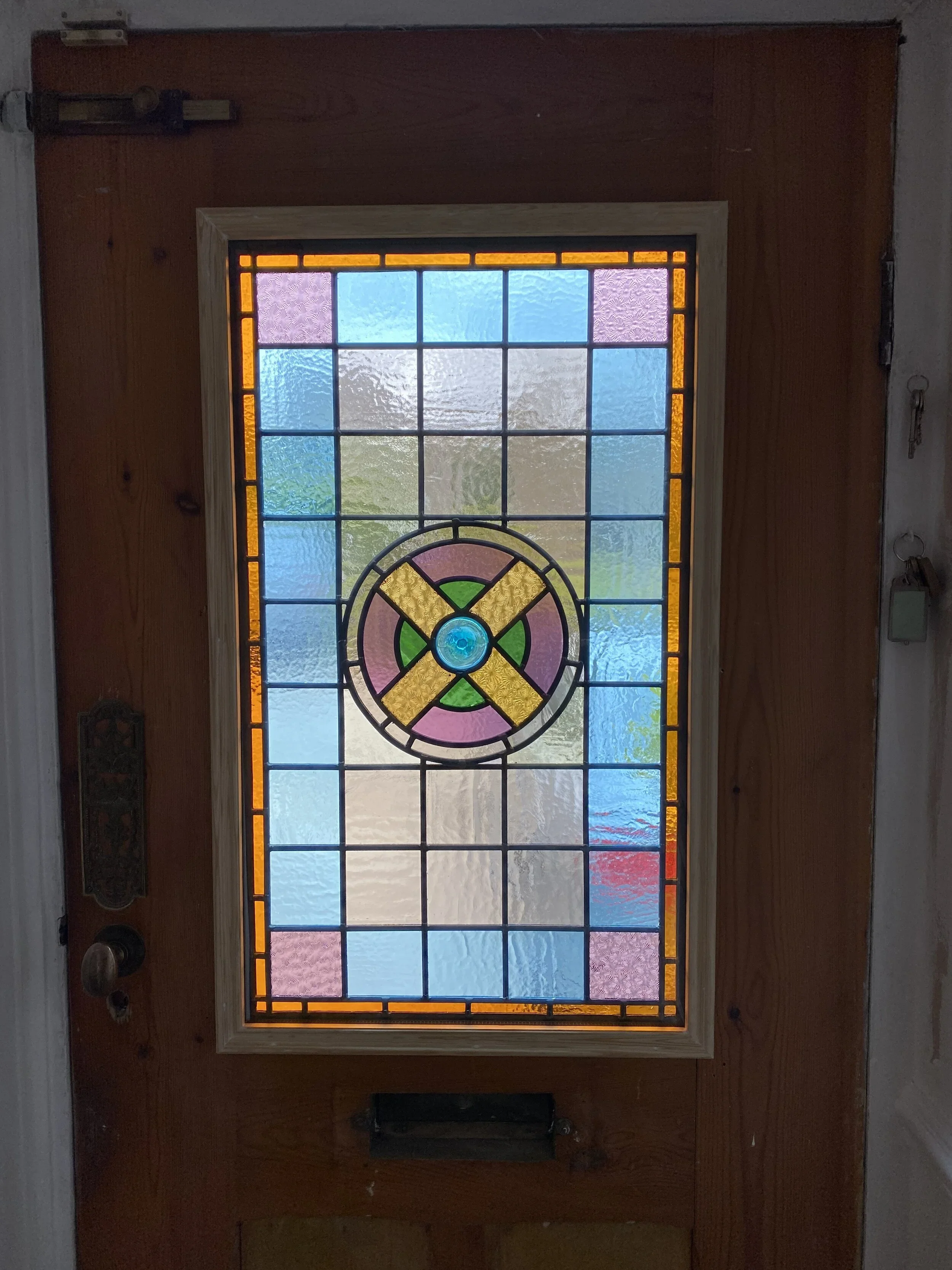 A wooden front door with a stained glass window featuring a circular pattern with pink, yellow, green, and blue glass sections, framed by colorful stained glass panels in pink, blue, and orange.