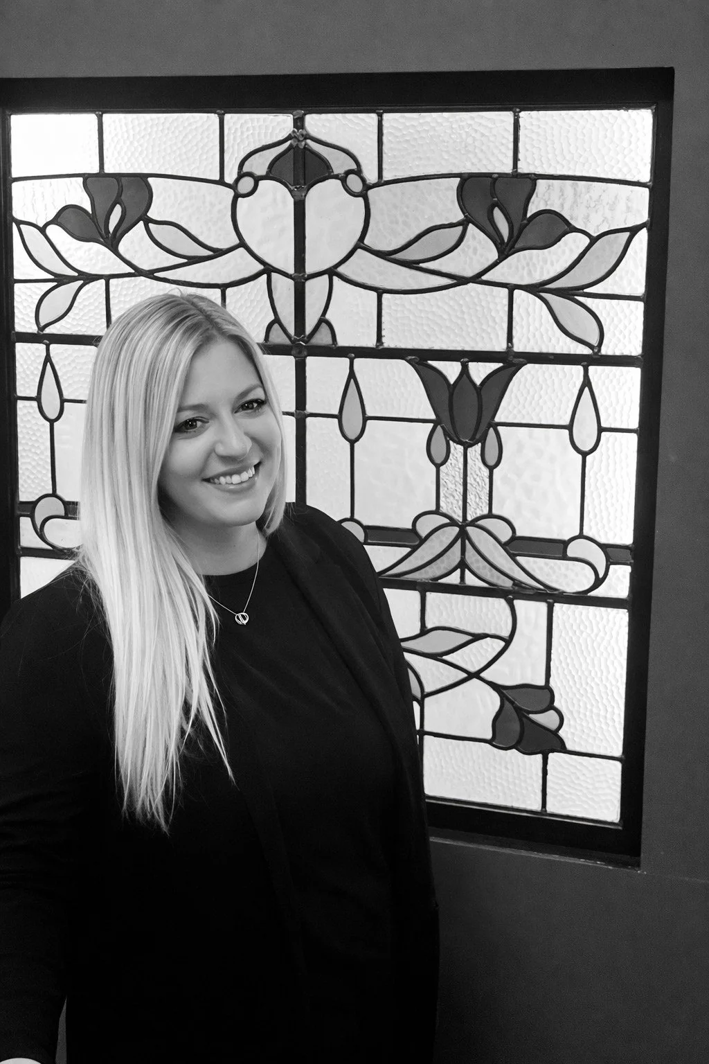 A woman with long blonde hair smiling, wearing a black blazer and a necklace, standing in front of a decorative stained glass window.