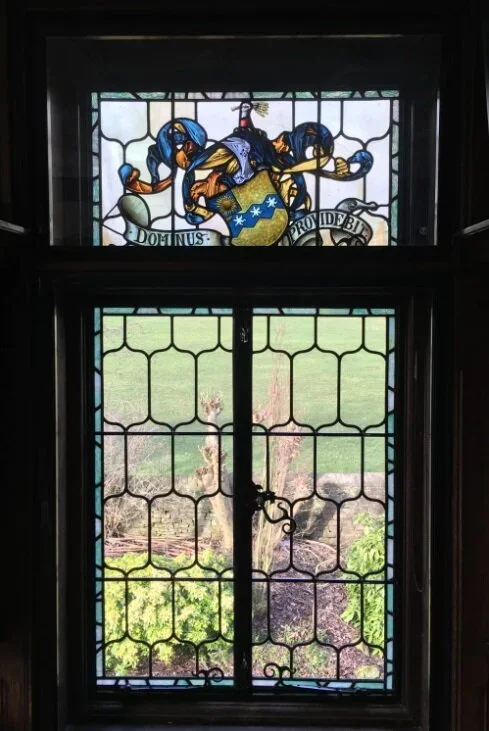 Stained glass window showing a heraldic shield with blue and gold decoration and a banner with the words 'DOMINUS PROVIDE EST', with greenery visible outside.
