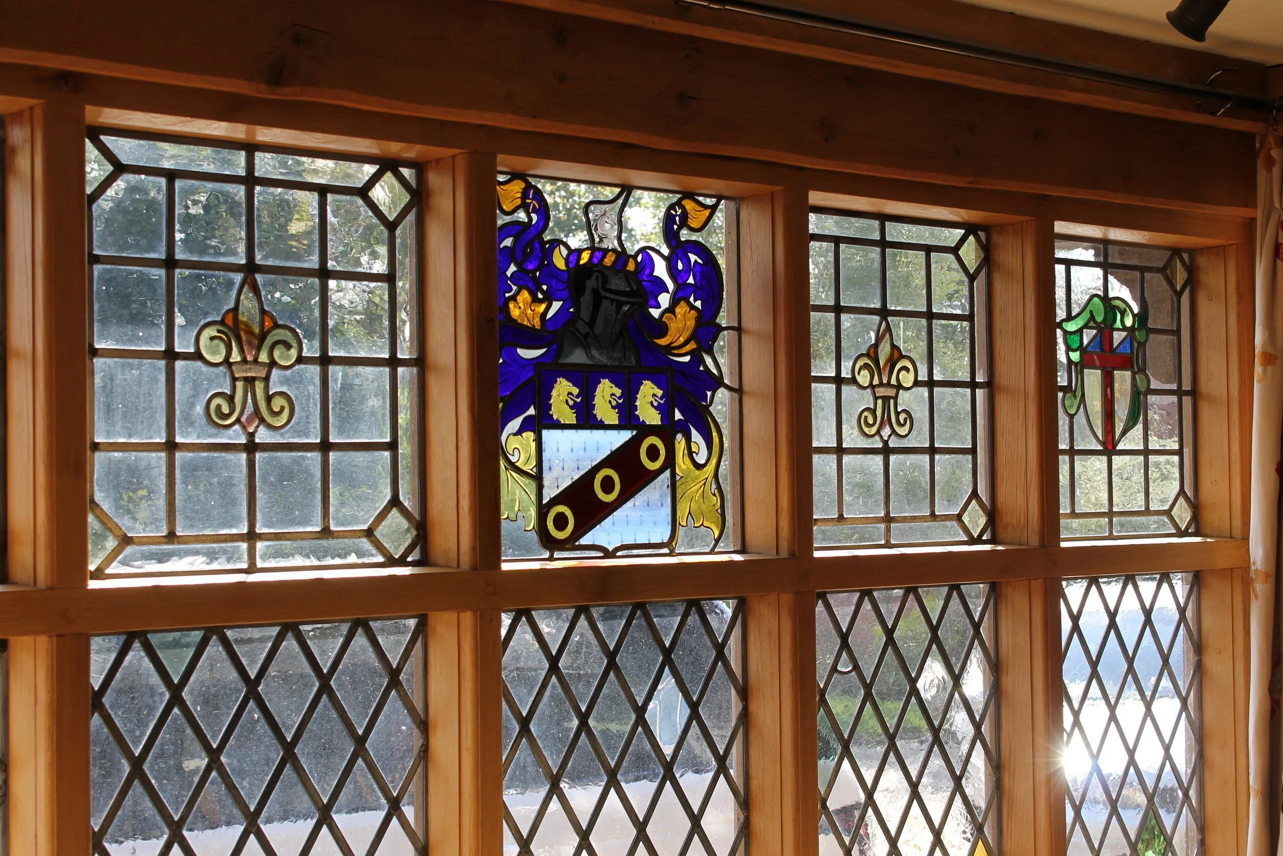 Decorative stained glass window with a coat of arms, flanked by two fleur-de-lis, set in a wooden frame with diamond-shaped glass panes, natural outdoor scenery visible through glass.