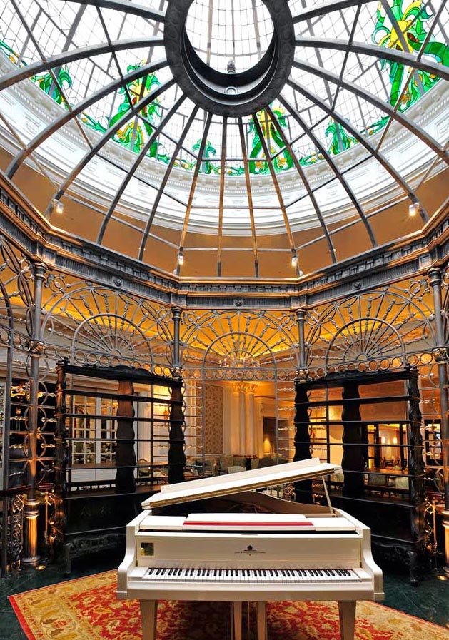 An elegant indoor space with a white grand piano on a red patterned rug, surrounded by ornate metal railings, under a glass-domed ceiling with decorative stained glass elements at the top.