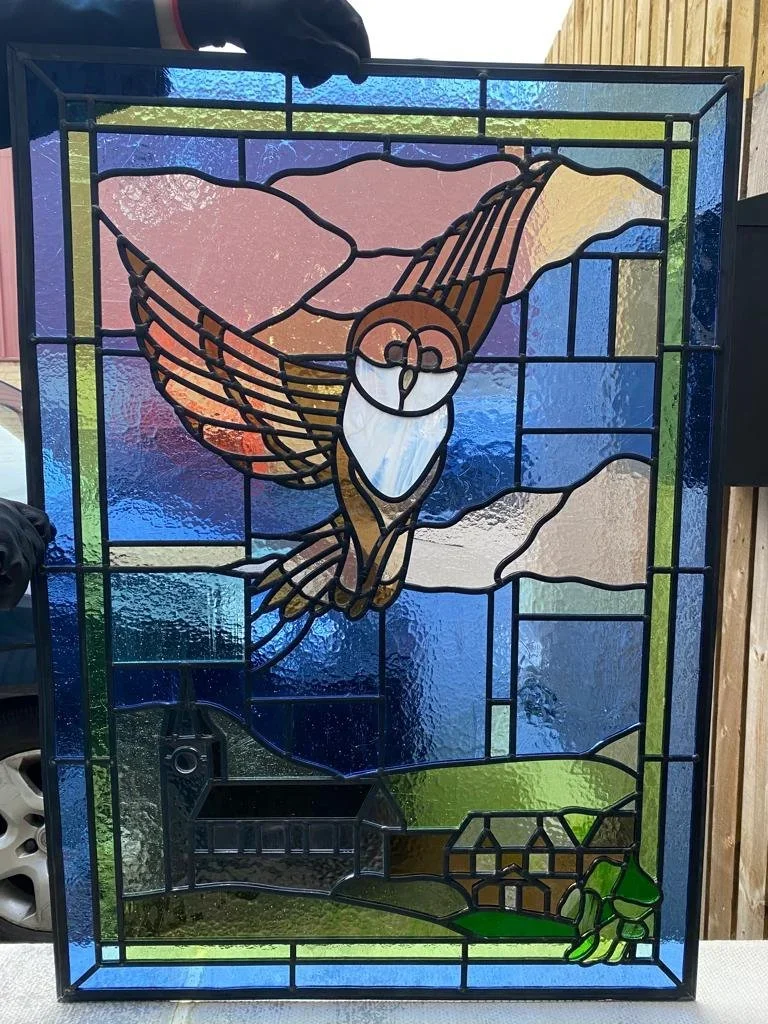 Colorful stained glass window featuring a bird in flight over a pastoral landscape with a house, trees, and mountains.