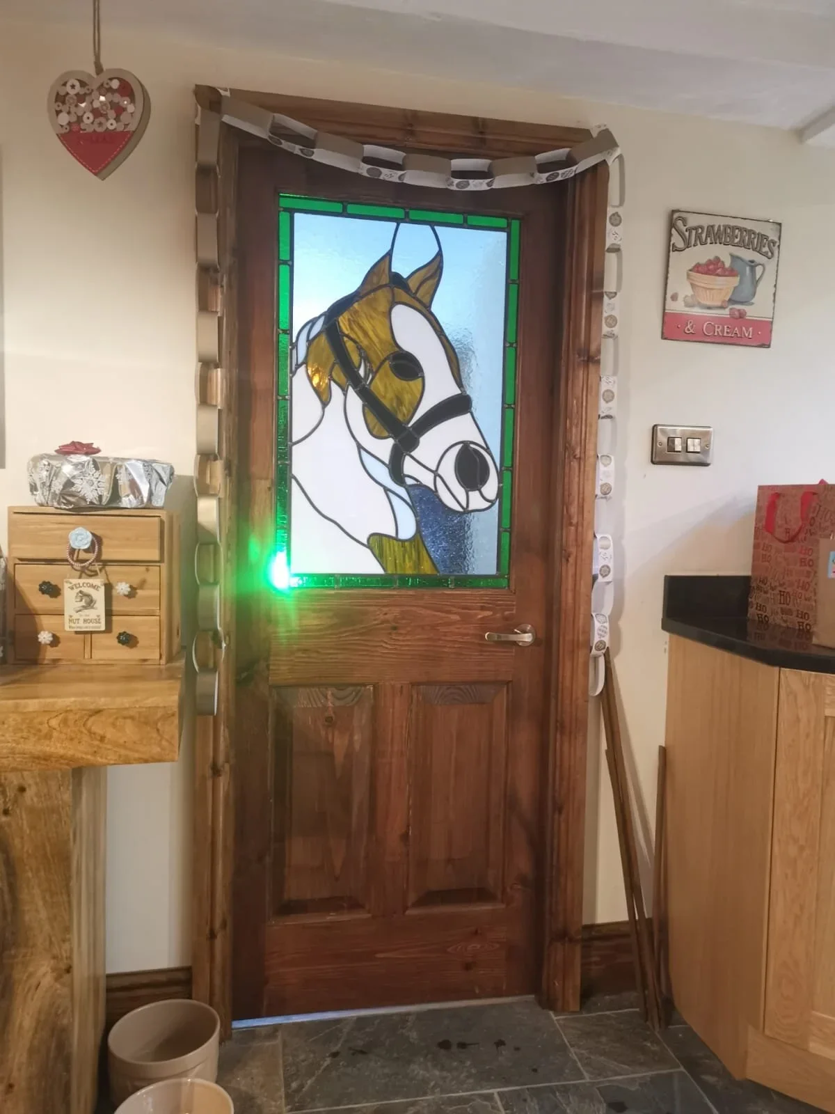 Decorated wooden door with a stained-glass panel depicting a horse's head, surrounded by a greenery border. The door is framed by a garland of white and gray paper rings. To the right, a vintage sign reads 'Strawberries & Cream,' and to the left, a s