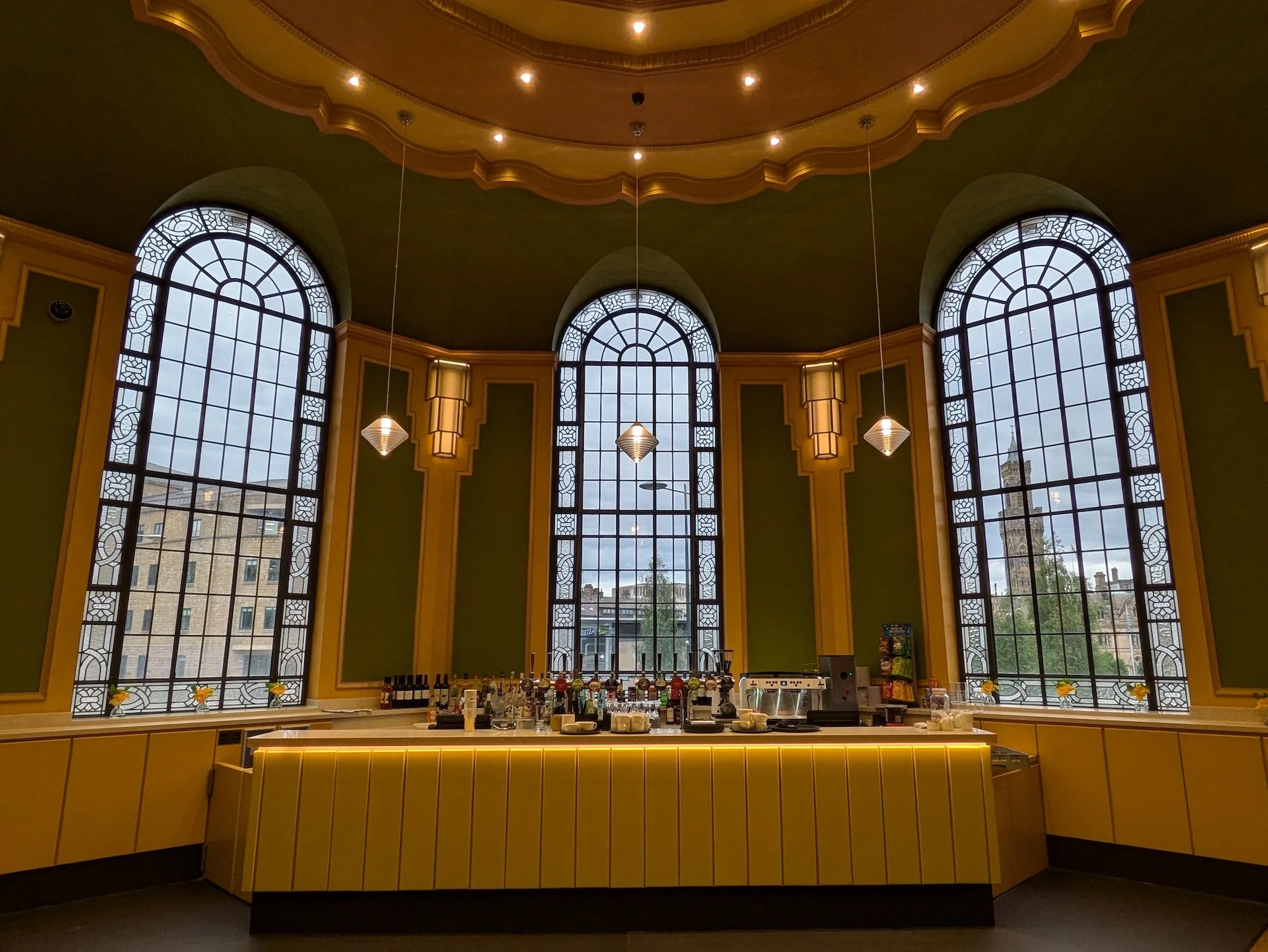 Interior of a cafe or restaurant with large arched windows, yellow walls, decorative lighting, and a bar counter with bottles and glasses.