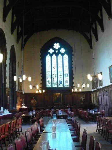 Interior of a historic dining hall with long wooden tables and chairs, tall stained glass window at the front, high ceiling with exposed dark wooden beams, and wall-mounted lights.