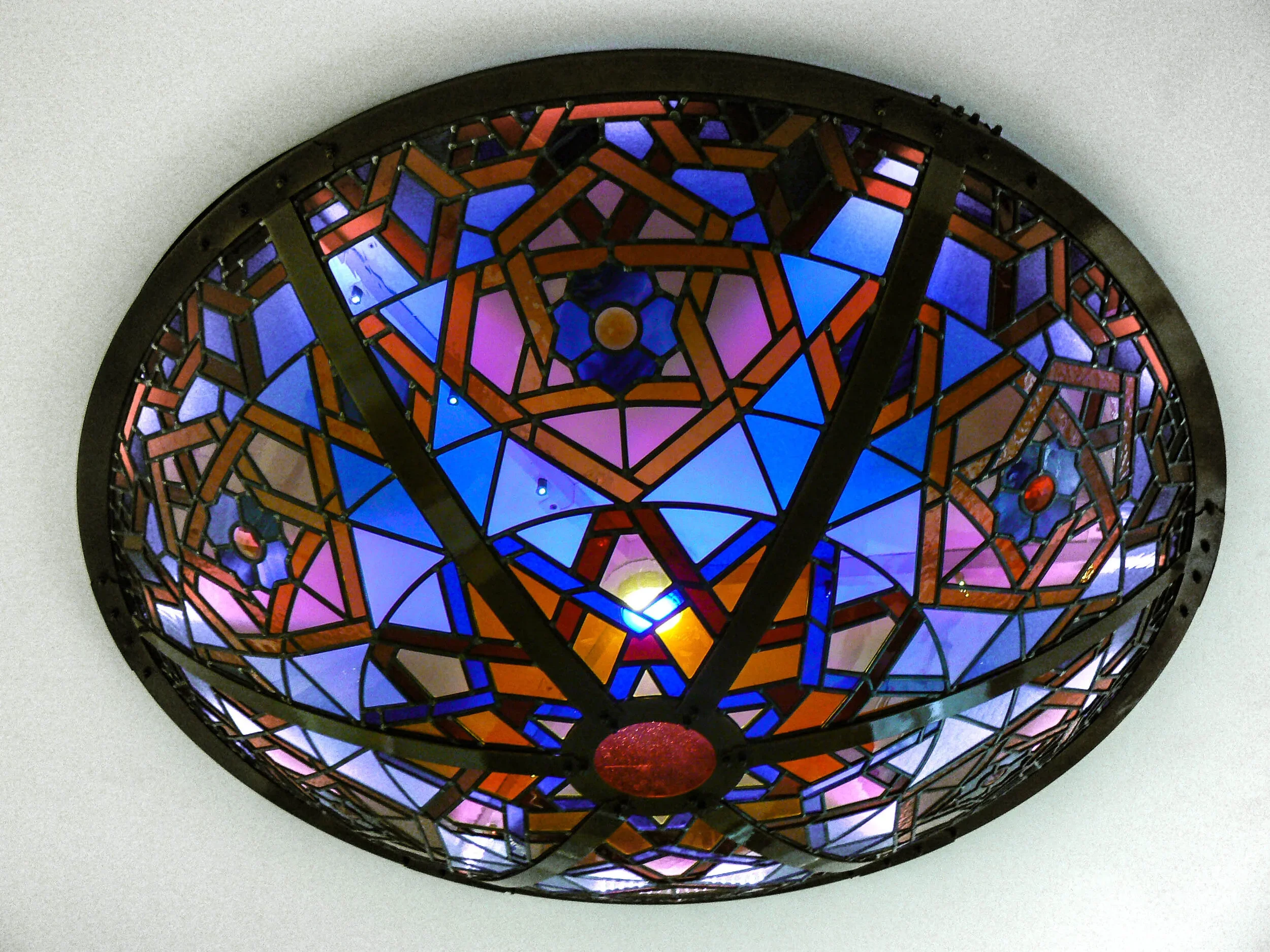 Colorful stained glass ceiling light fixture with geometric patterns, illuminated from below, set against a white ceiling.