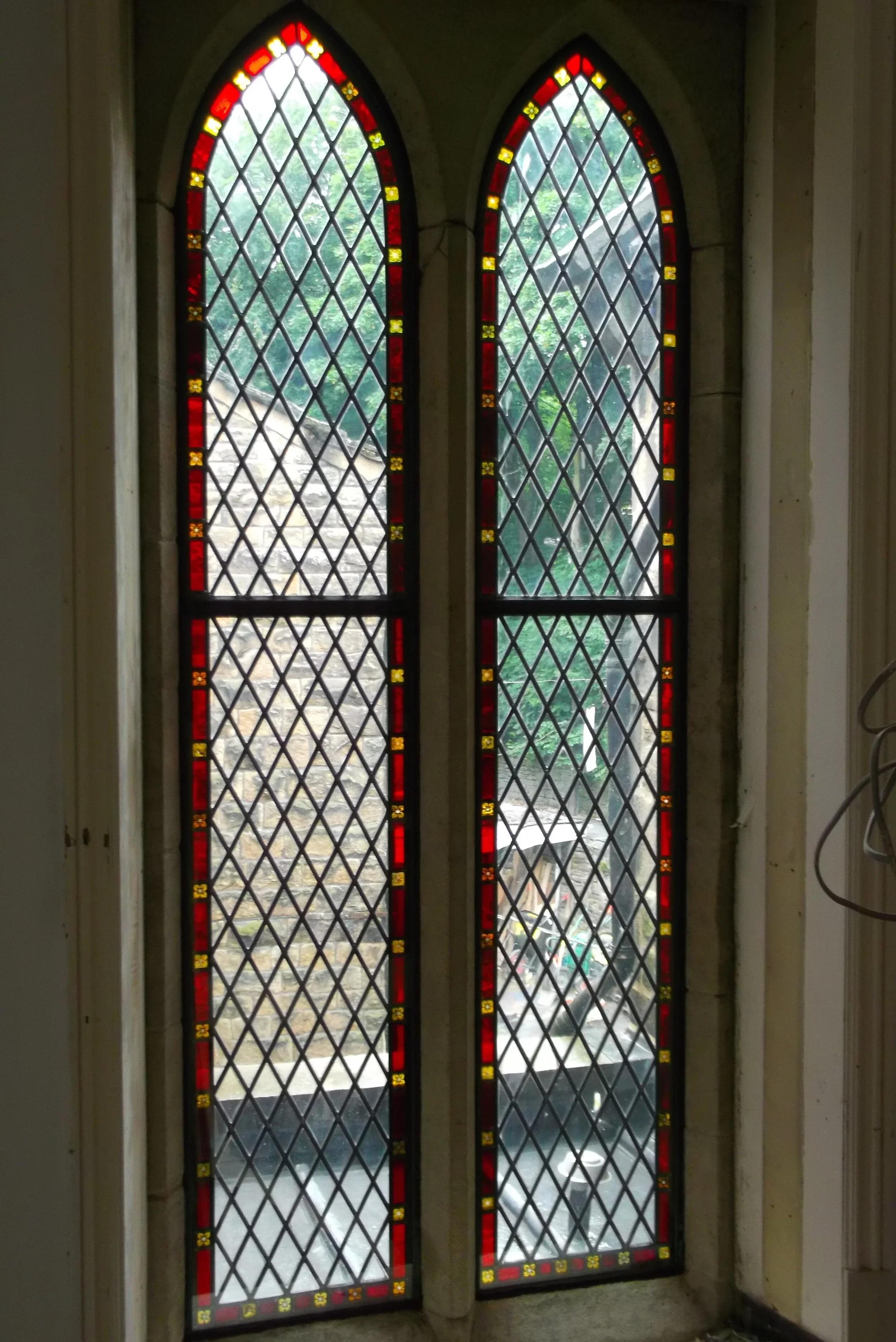 Two tall stained glass windows with diamond patterns and red and yellow border accents, set in a stone frame, with trees visible outside.