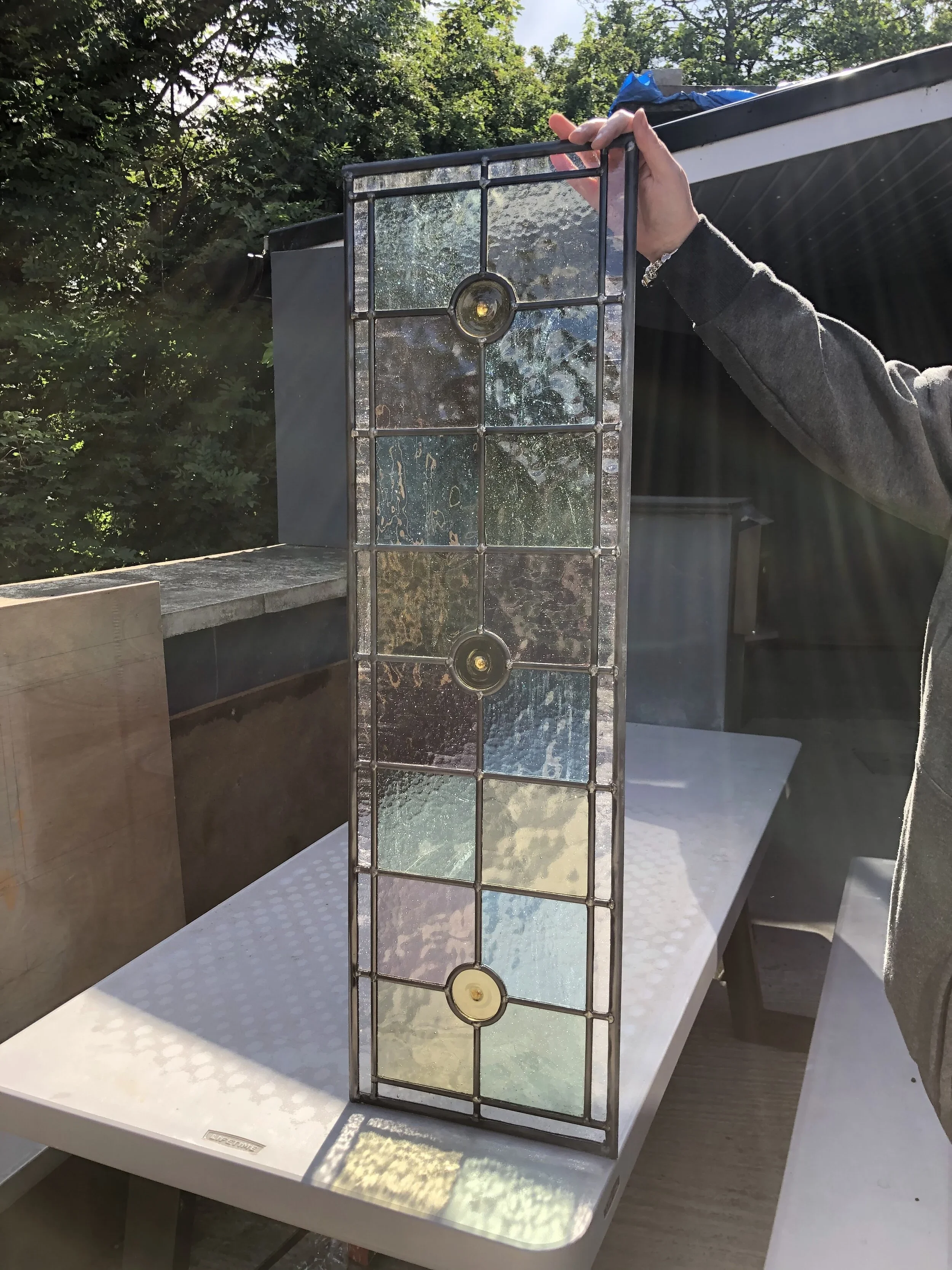 A person holding a stained glass window panel with a circular design in the center, outdoors with trees and sunlight.