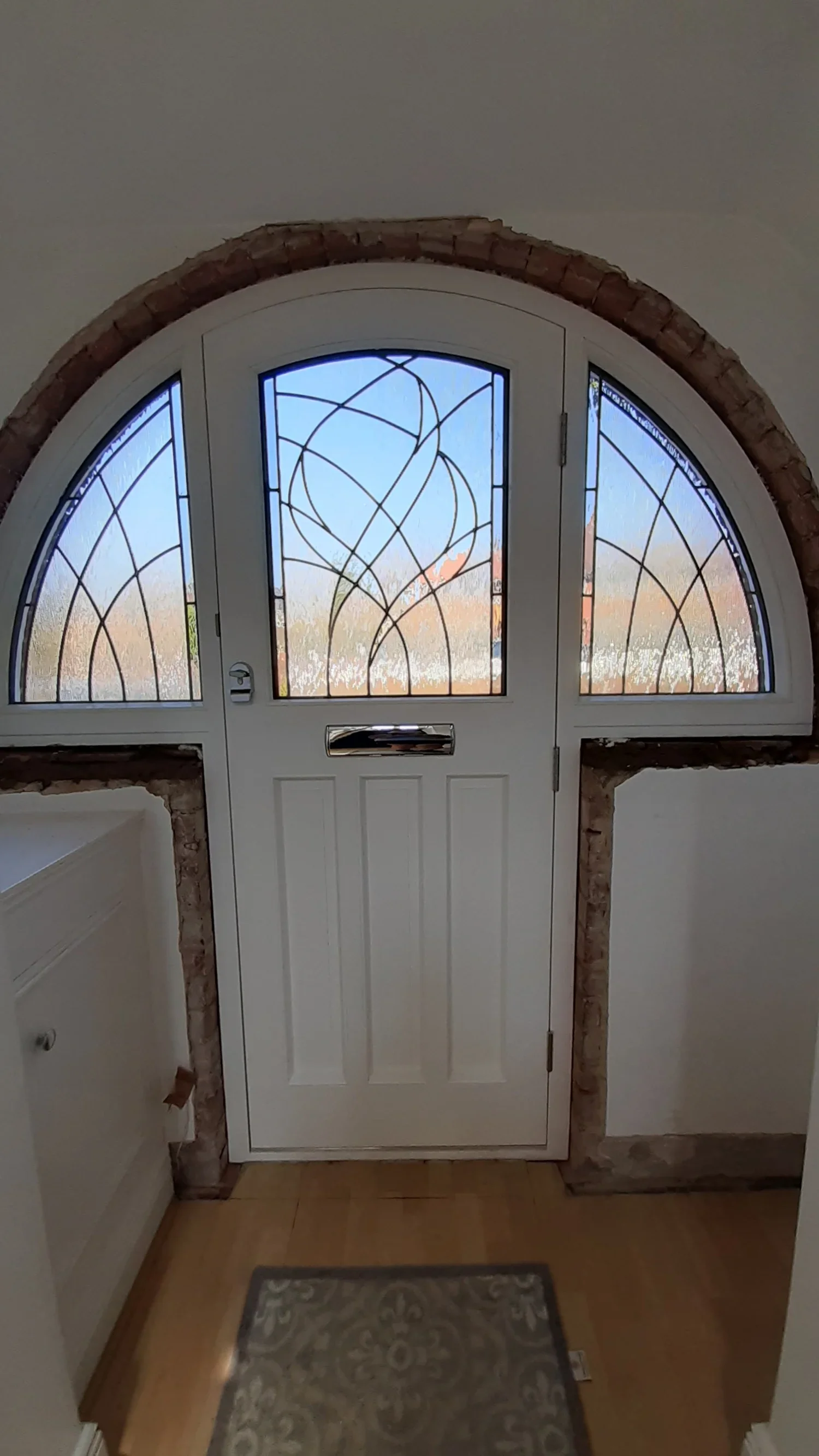 Interior view of a white front door with decorative glass panels, surrounded by brick archway, and a small rug in front.