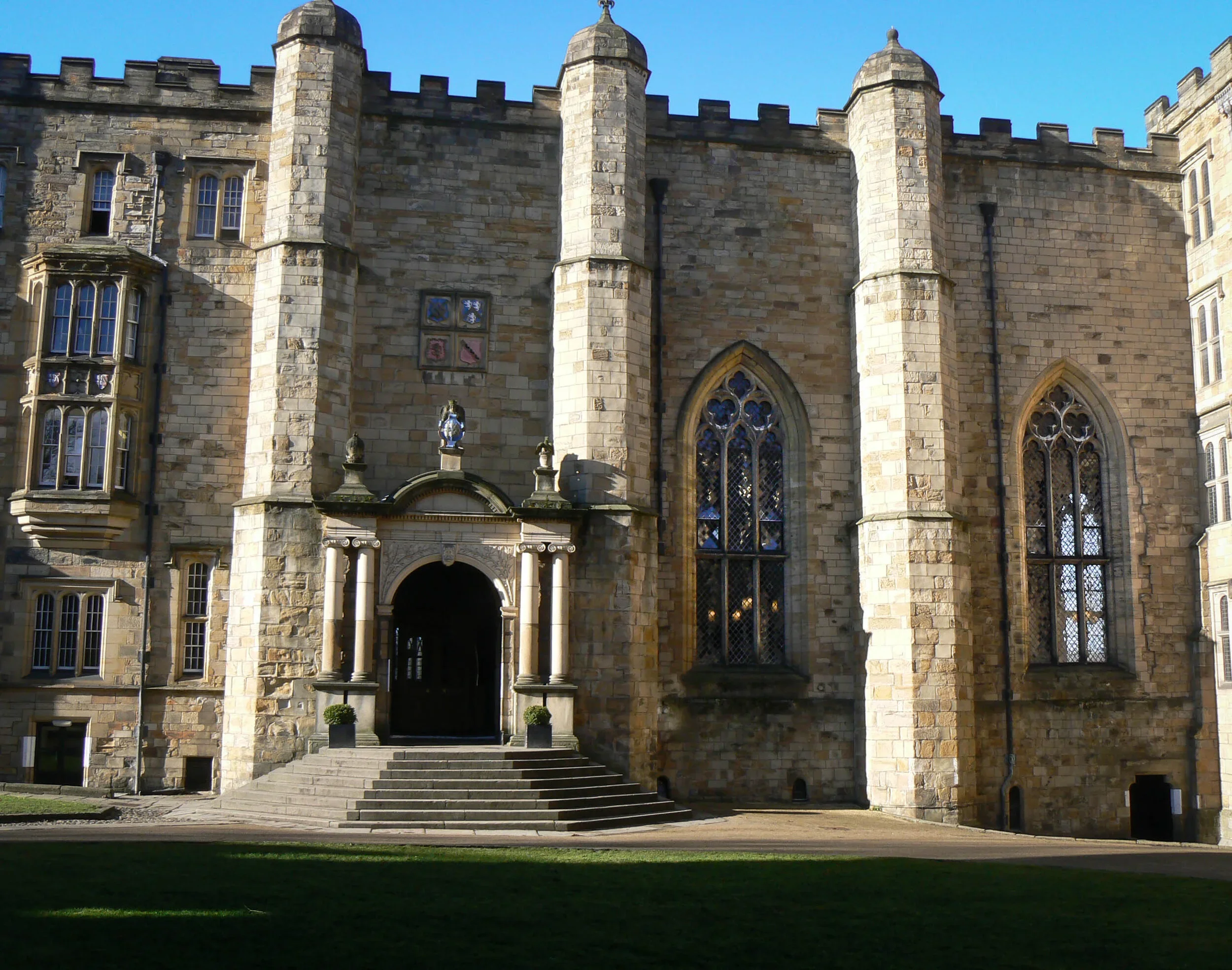 A stone castle or mansion with Gothic architecture, featuring large arched windows, a turret, and a staircase leading to an entrance, under a clear blue sky.