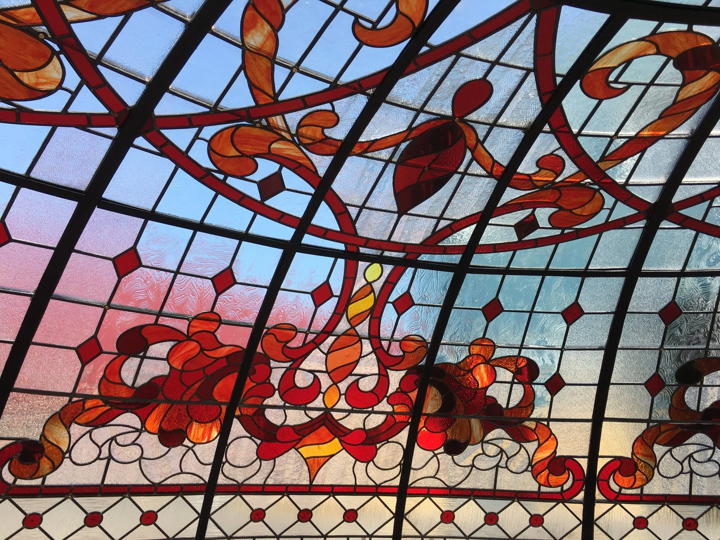 View of a stained glass ceiling featuring red, orange, and yellow swirling patterns with black borders, through which blue sky with clouds is visible.