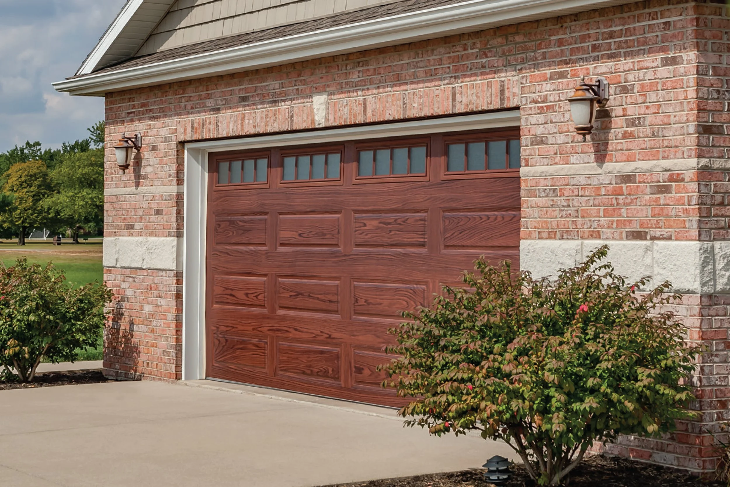 Red wooden garage door on a brick house with outdoor wall-mounted lights and bushes in front