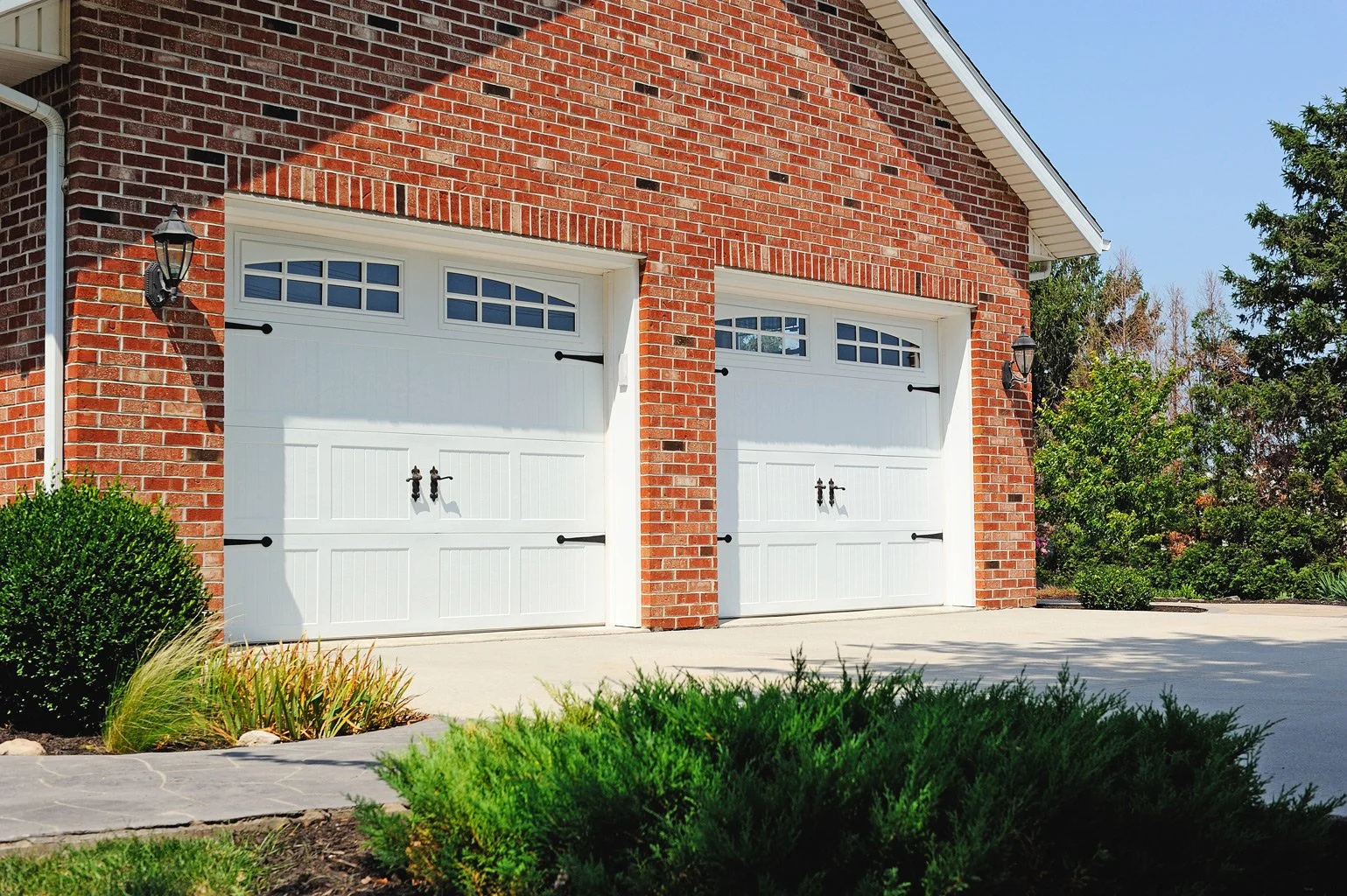 Two white garage doors with windows at the top, attached to a red brick house, surrounded by shrubs and trees, on a concrete driveway under a clear blue sky.