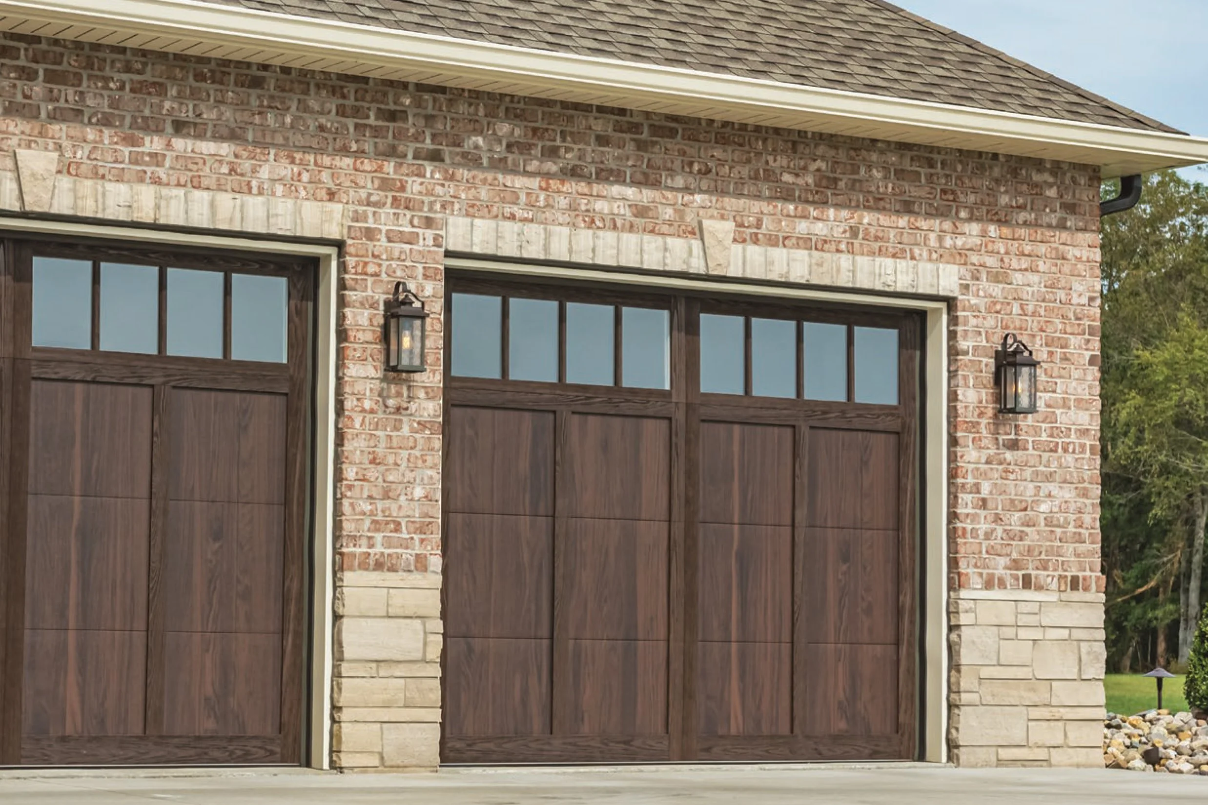 A garage with two wooden doors and brick exterior, featuring black lantern-style lights on each side, and a stone accent at the bottom of the brick wall.