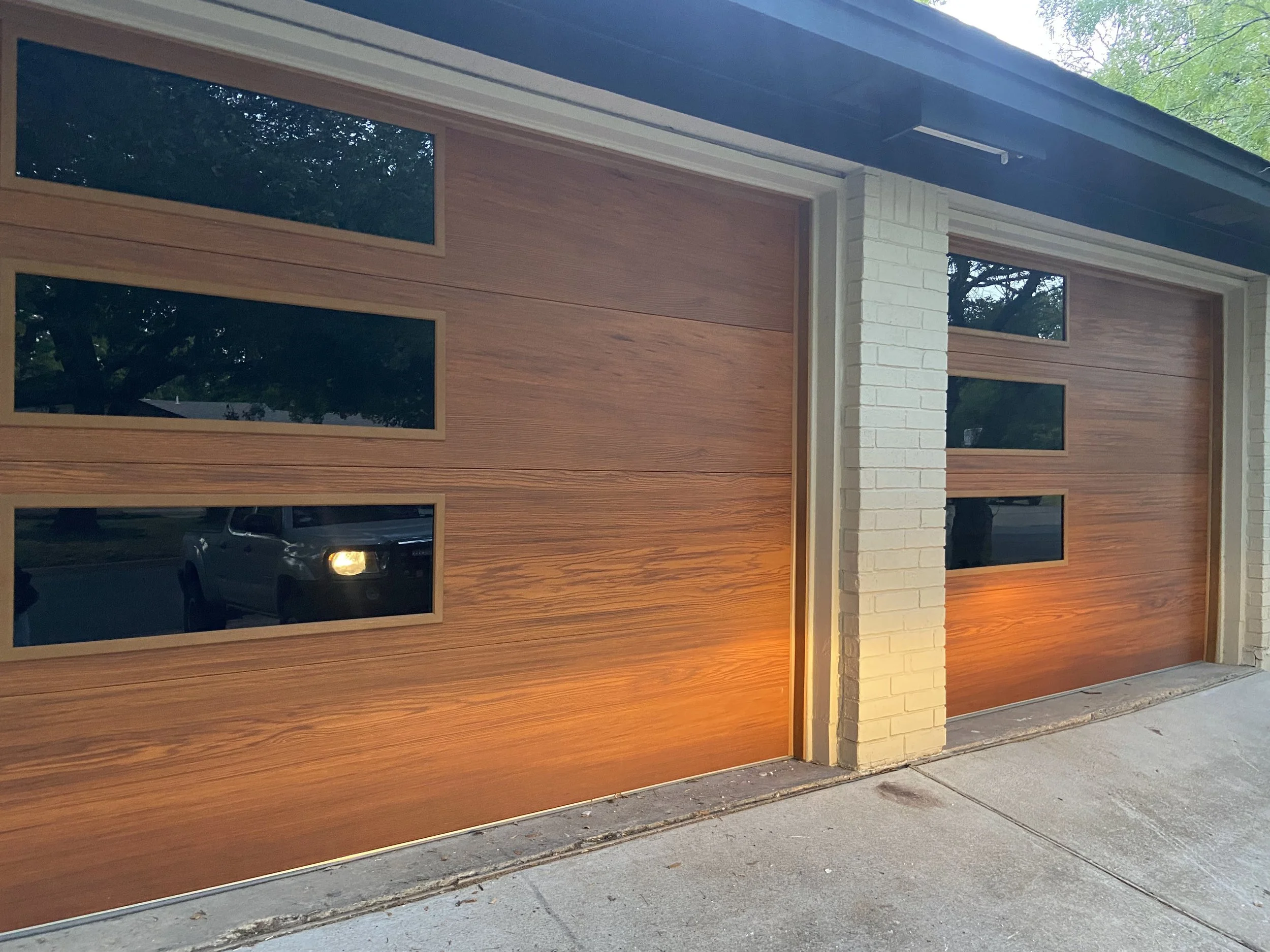 Two modern garage doors with wood paneling and horizontal rectangular windows, adjacent to white brick columns, with small outdoor lights at the base and a driveway in front.
