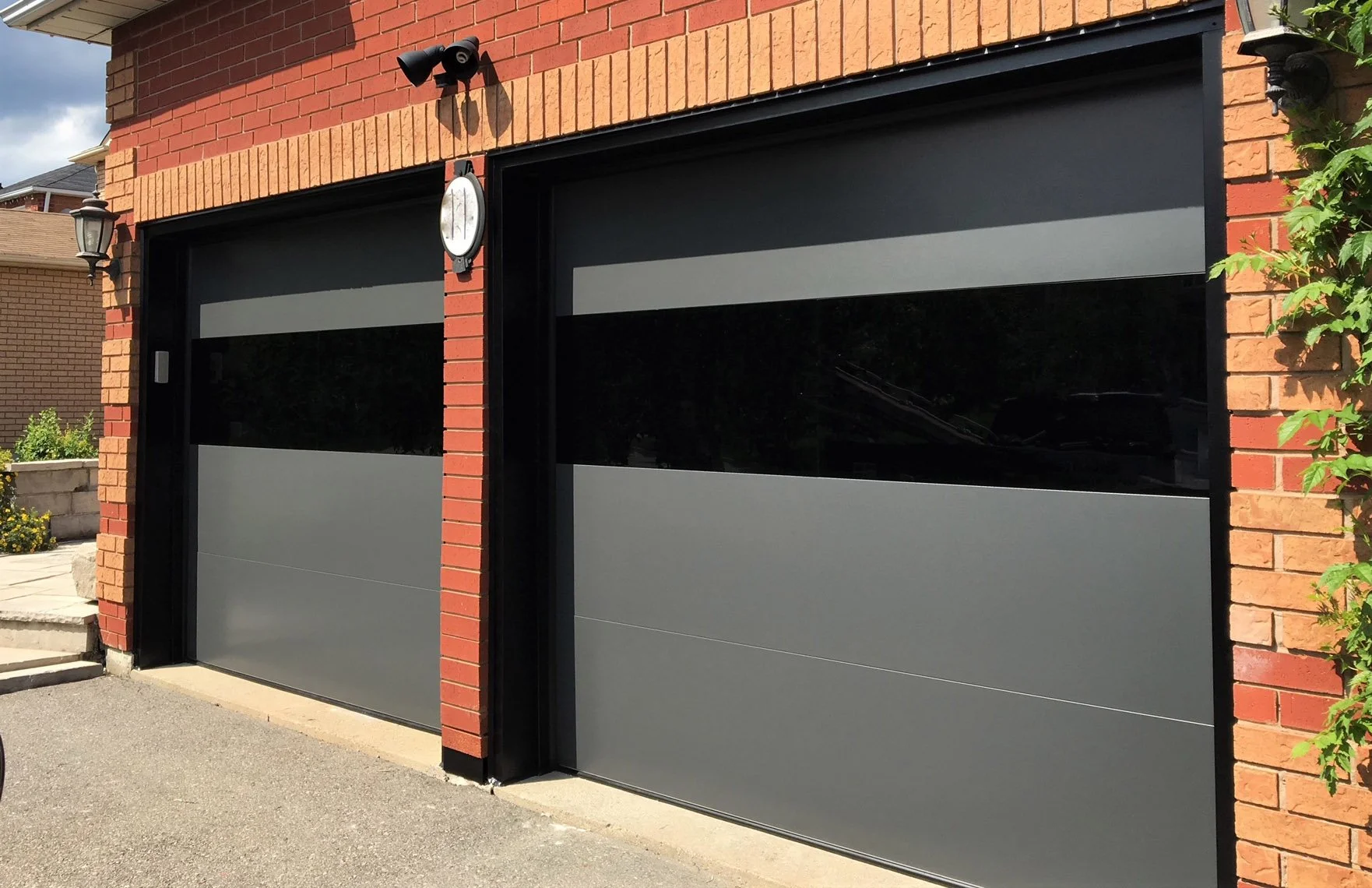 Two modern gray garage doors with black frames and a black track, attached to a red brick house. A small outdoor wall lamp and a round clock are mounted above the left garage door. A small part of a bicycle wheel is visible in the bottom left corner, and some greenery is seen on the right side of the image.