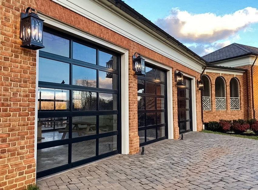 A building with three large black-framed garage doors, brick walls, decorative black outdoor lanterns, and a paved stone patio. Modern windows and a section with arched windows and a small decorative balcony are visible.