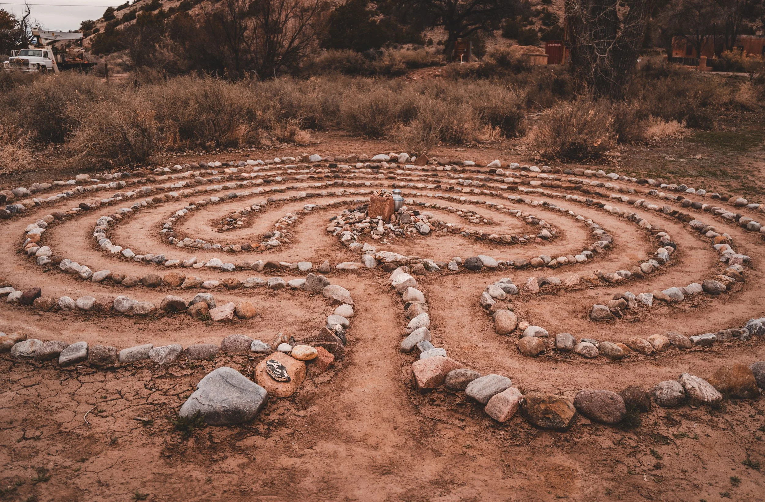 A spiral labyrinth made of rocks on a dirt ground in a desert landscape, with dry bushes and sparse trees in the background.