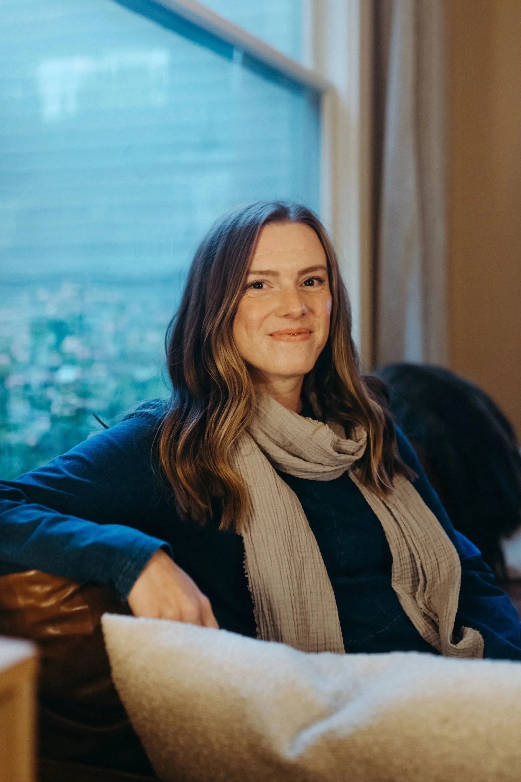 A woman with long wavy brown hair, wearing a beige scarf and a dark blue jacket, sitting by a window in a cozy indoor setting.