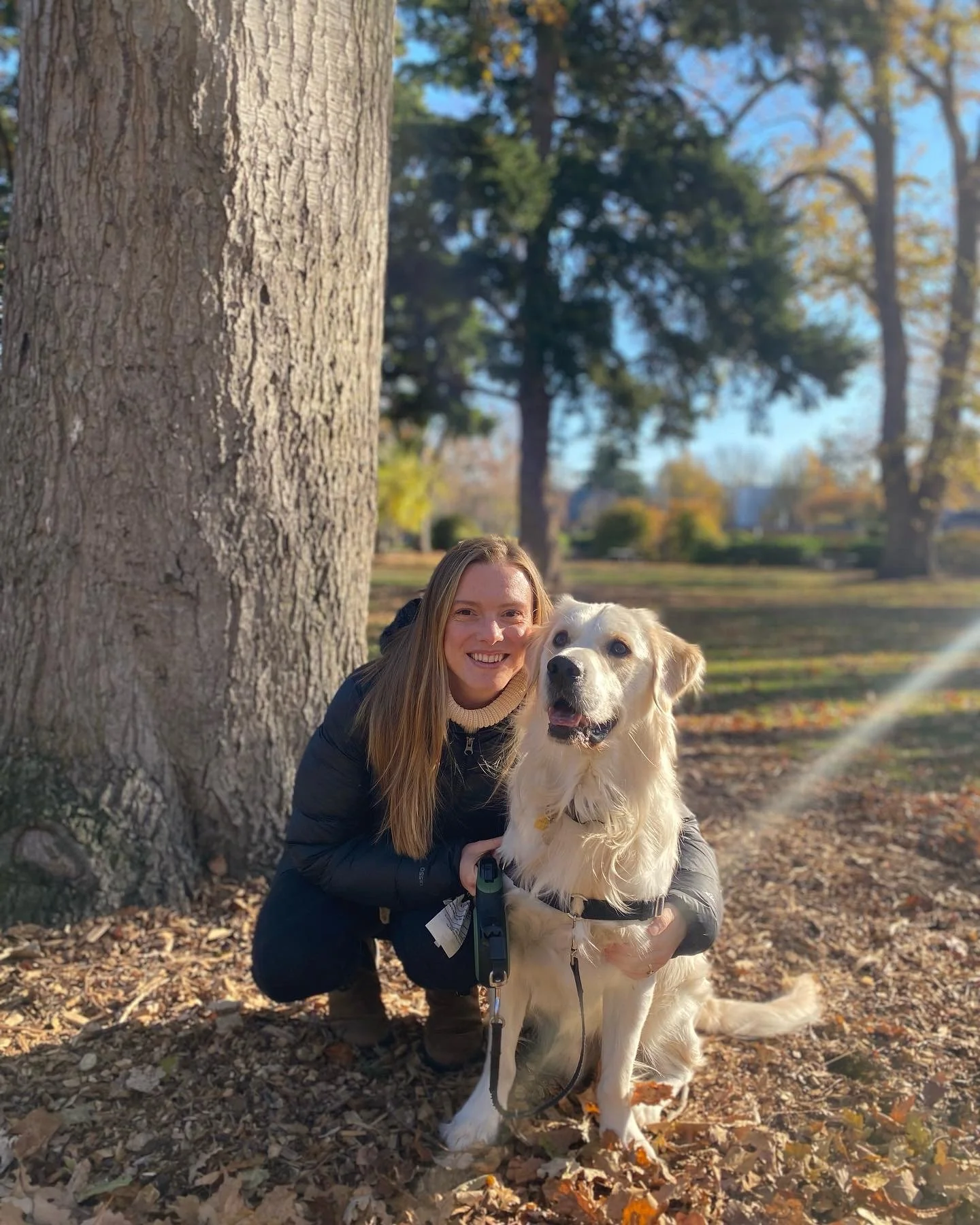 A woman with long blonde hair smiling and squatting beside a golden retriever dog in a park with large trees, fallen leaves, and blue sky in the background.