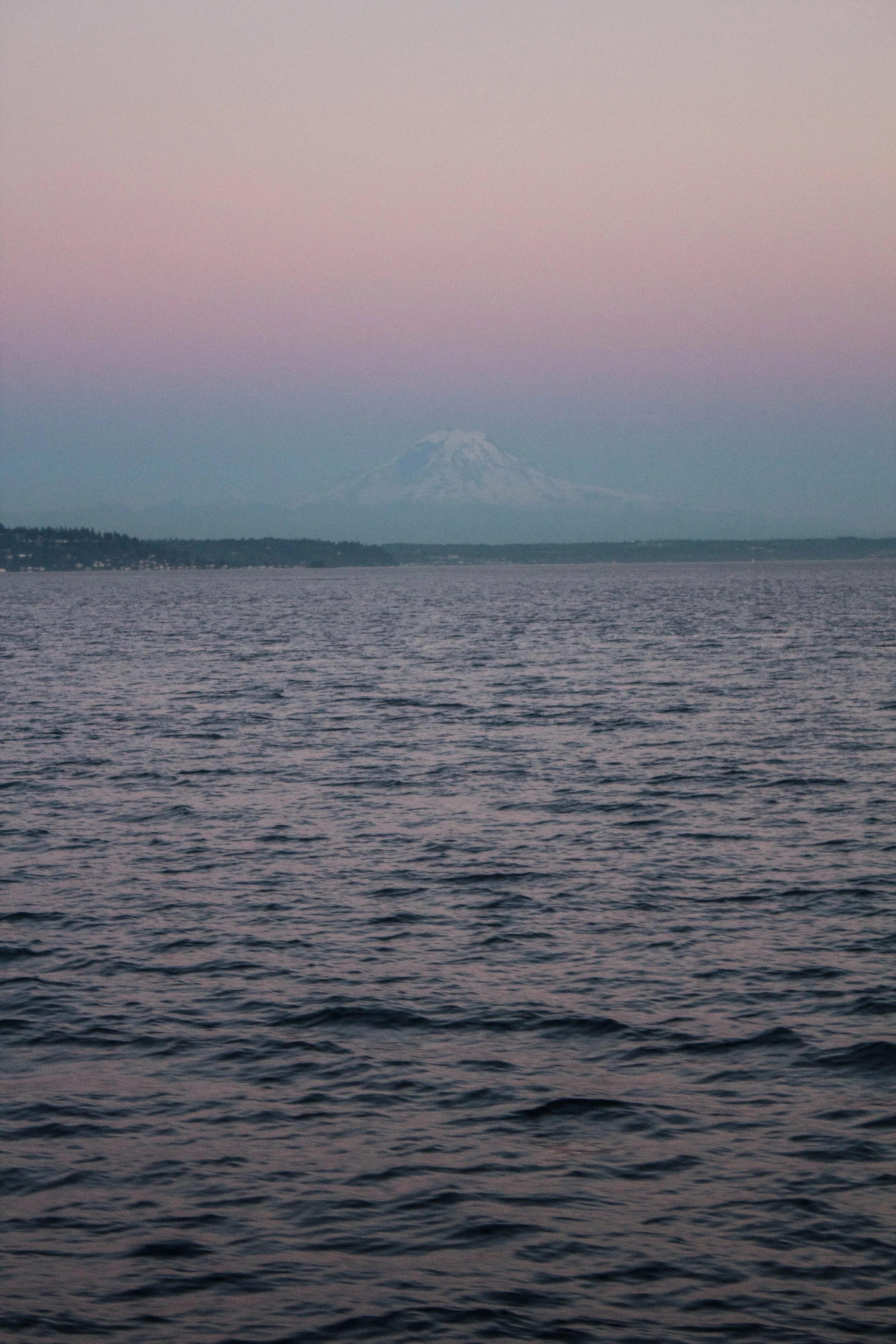 View of the ocean with small waves and a mountain in the background at sunset, with a pastel-colored sky.