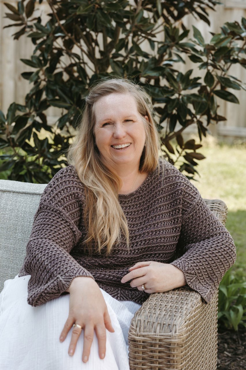 A woman with long blonde hair smiling while sitting outdoors on a wicker chair, with a large leafy plant in the background.