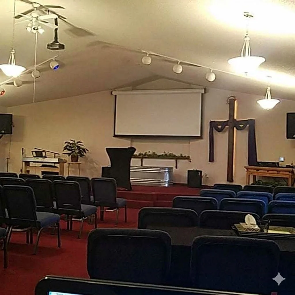 Inside a church sanctuary with rows of black chairs, a cross decorated with black fabric, a large projection screen, and a pulpit on a small stage, with ceiling lights and a ceiling fan.