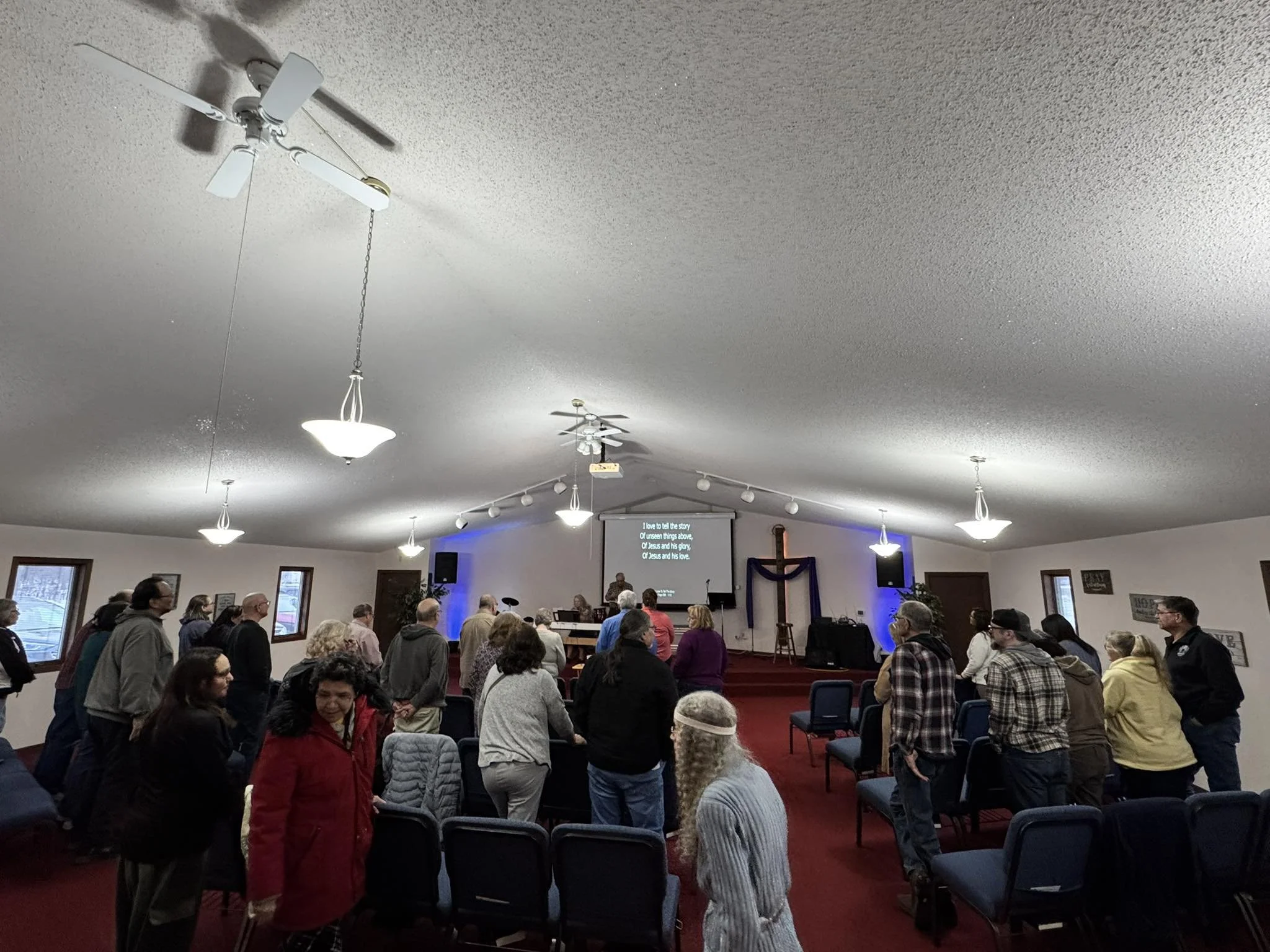 People gather and participate in a church service or worship in a room with a stage, a cross at the front, and a screen with song lyrics.