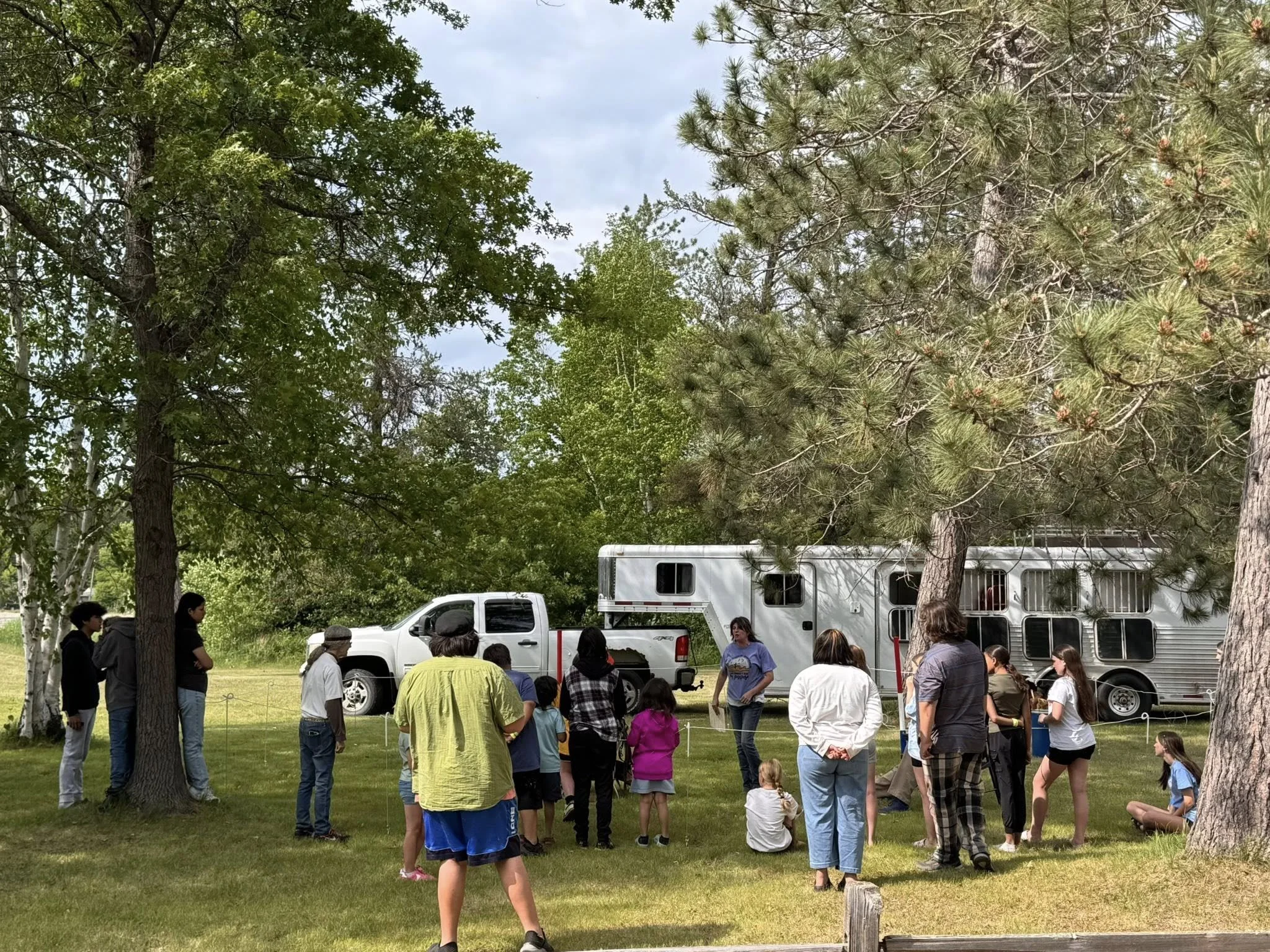 A group of people, including children and adults, gathered outdoors on a grassy area with trees, with a white truck and a horse trailer in the background.