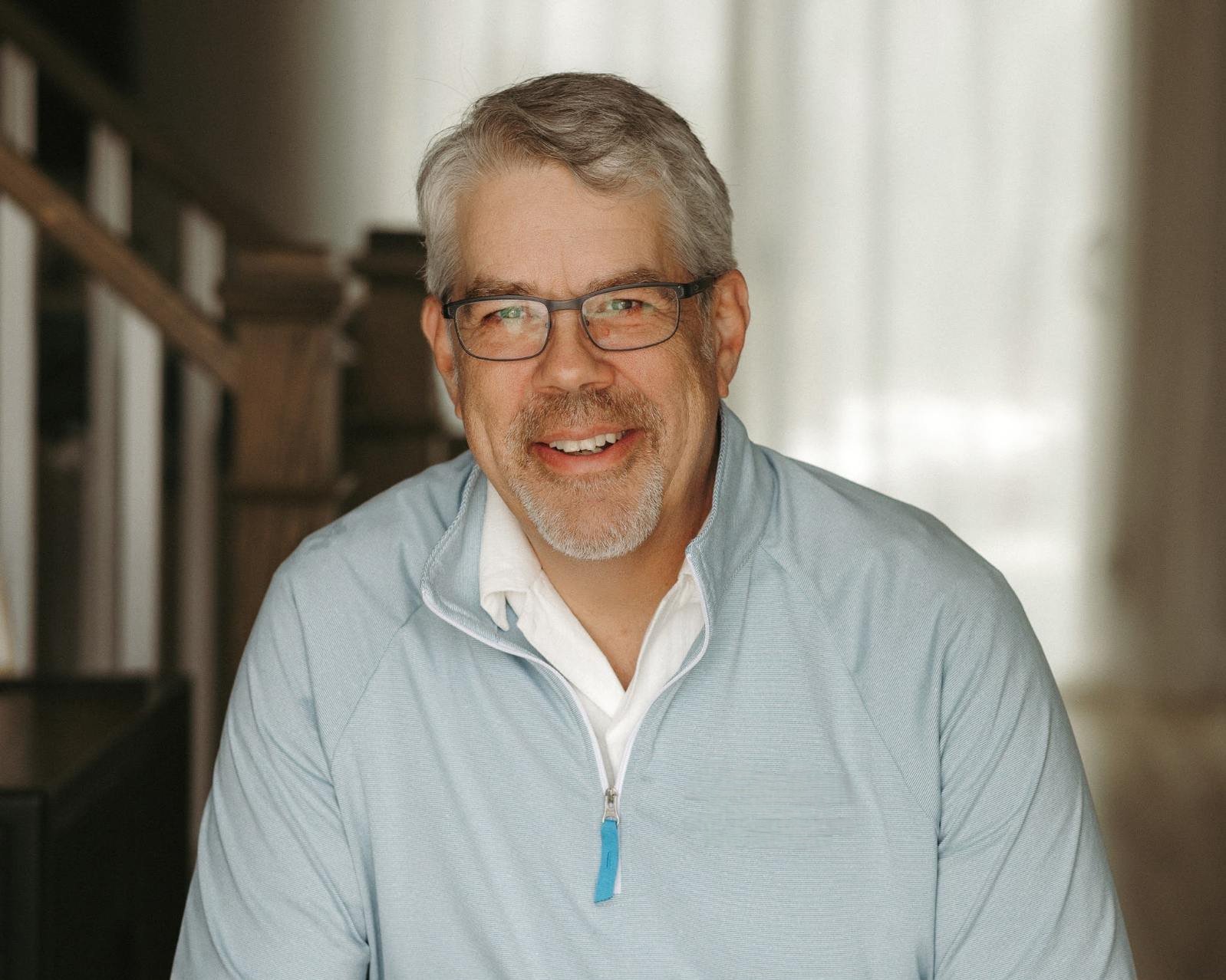 A smiling older man with glasses, gray hair, and a beard, wearing a light blue jacket and white shirt indoors.