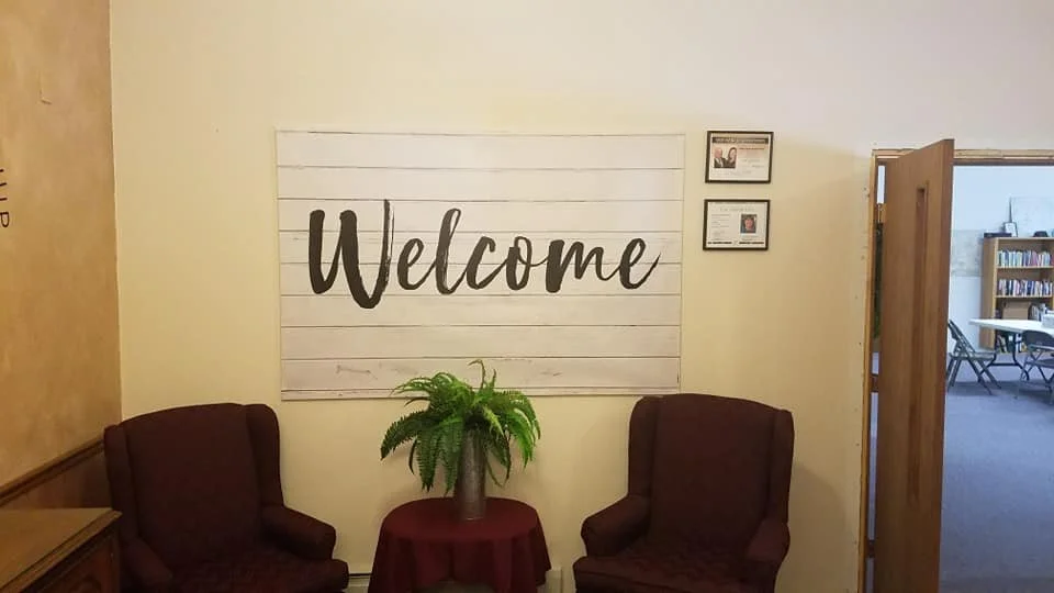 A welcoming interior with a white sign that says 'Welcome', two chairs, a small table with a potted fern, and a door leading to a room with bookshelves and tables.