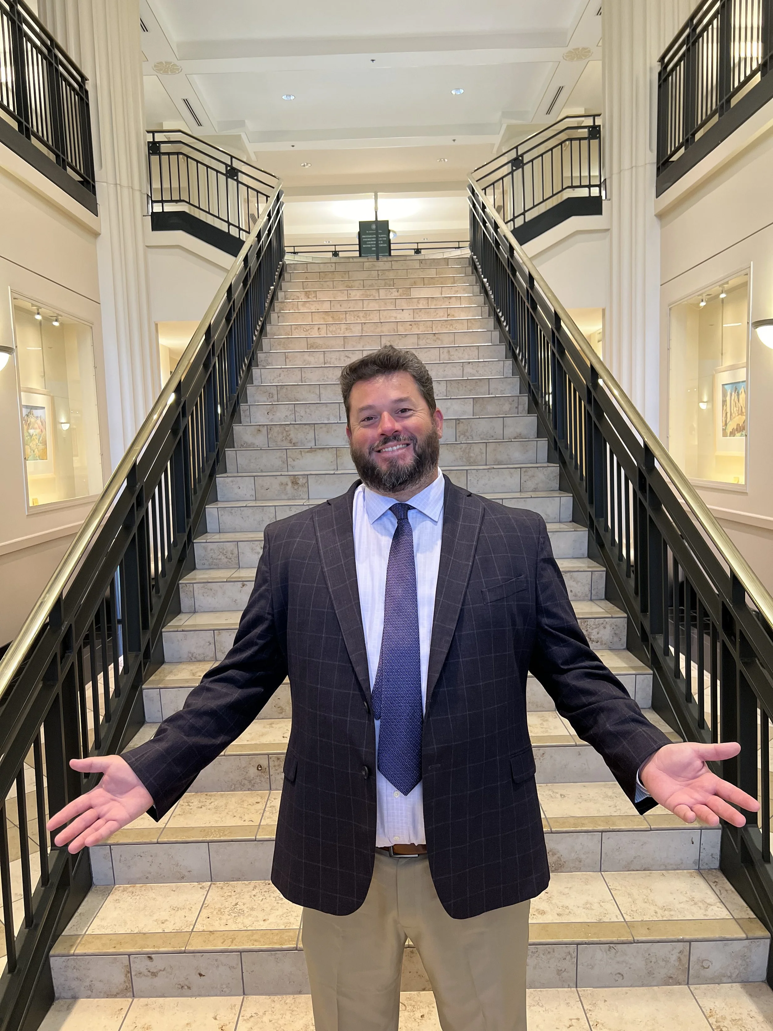 A man with a beard in a business suit stands at the bottom of a staircase, with his arms open wide, inside a building with framed artwork on the walls and a high ceiling.