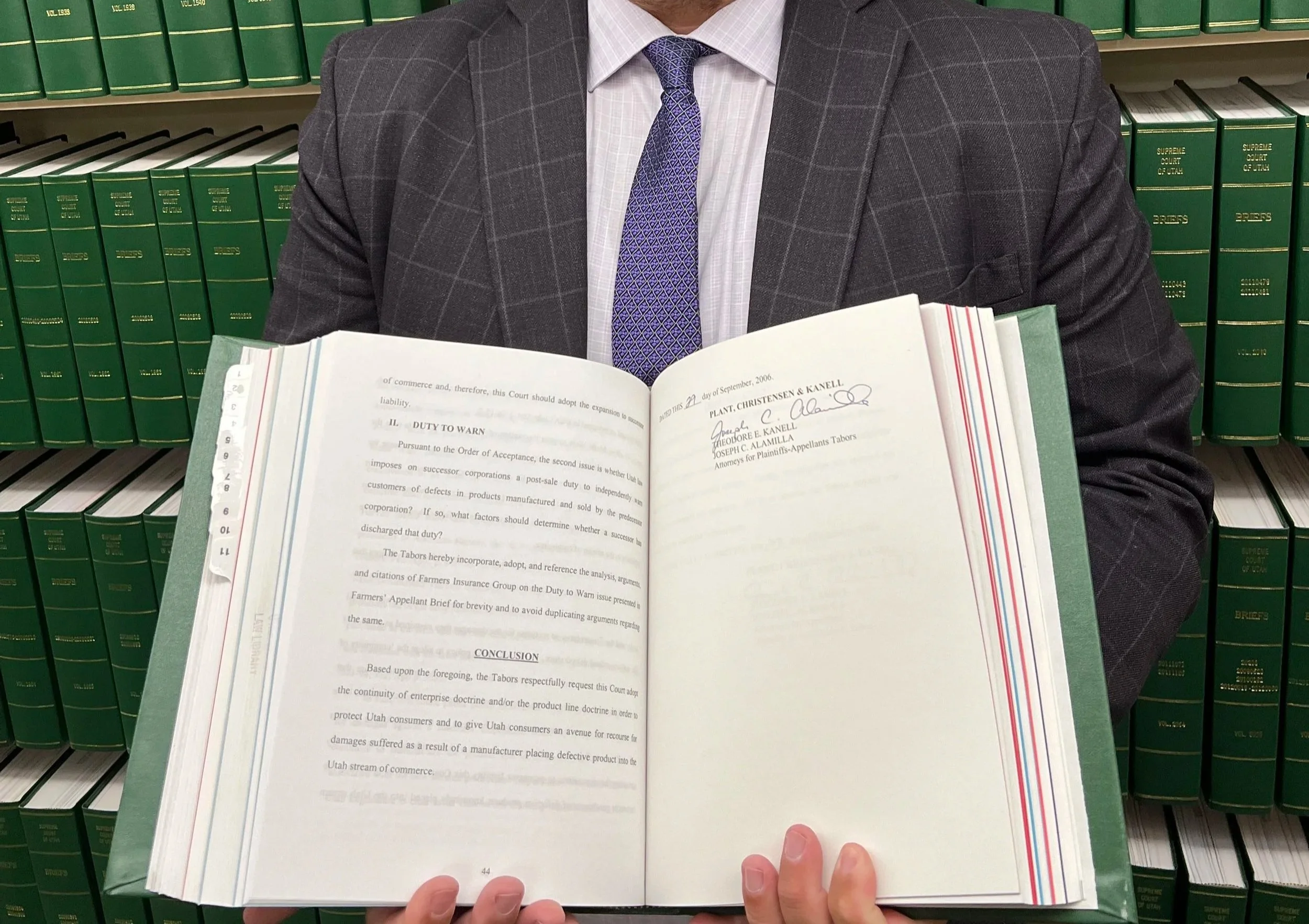 A person in a suit holding an open legal book in front of a background of green legal bookshelves.