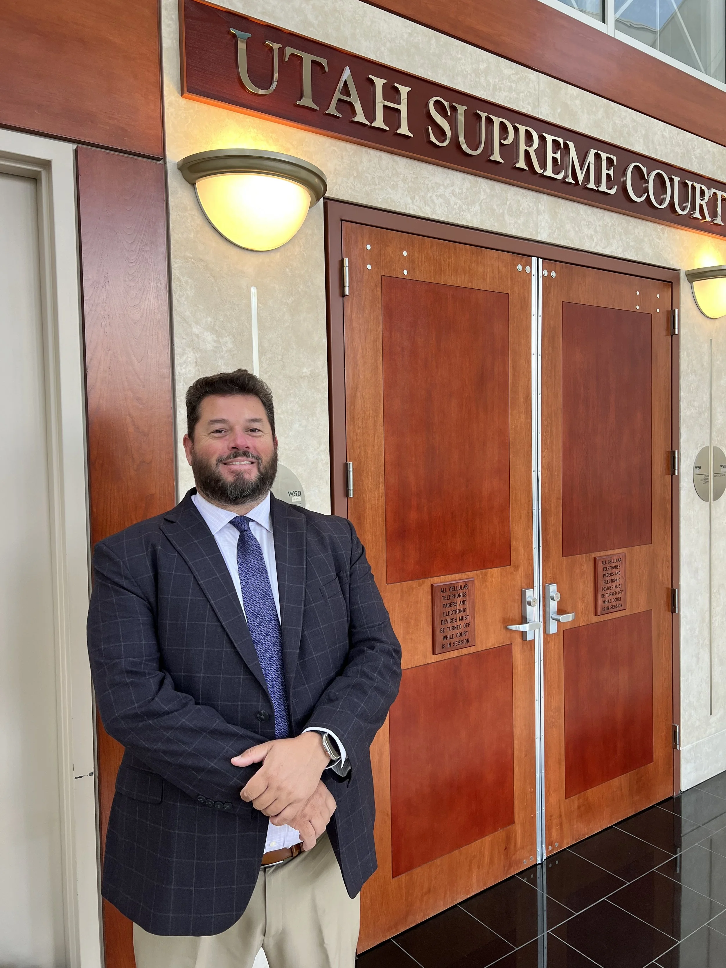 A man wearing a dark suit with a blue checkered shirt and tie, standing with his arms crossed, smiling in front of the Utah Supreme Court entrance.