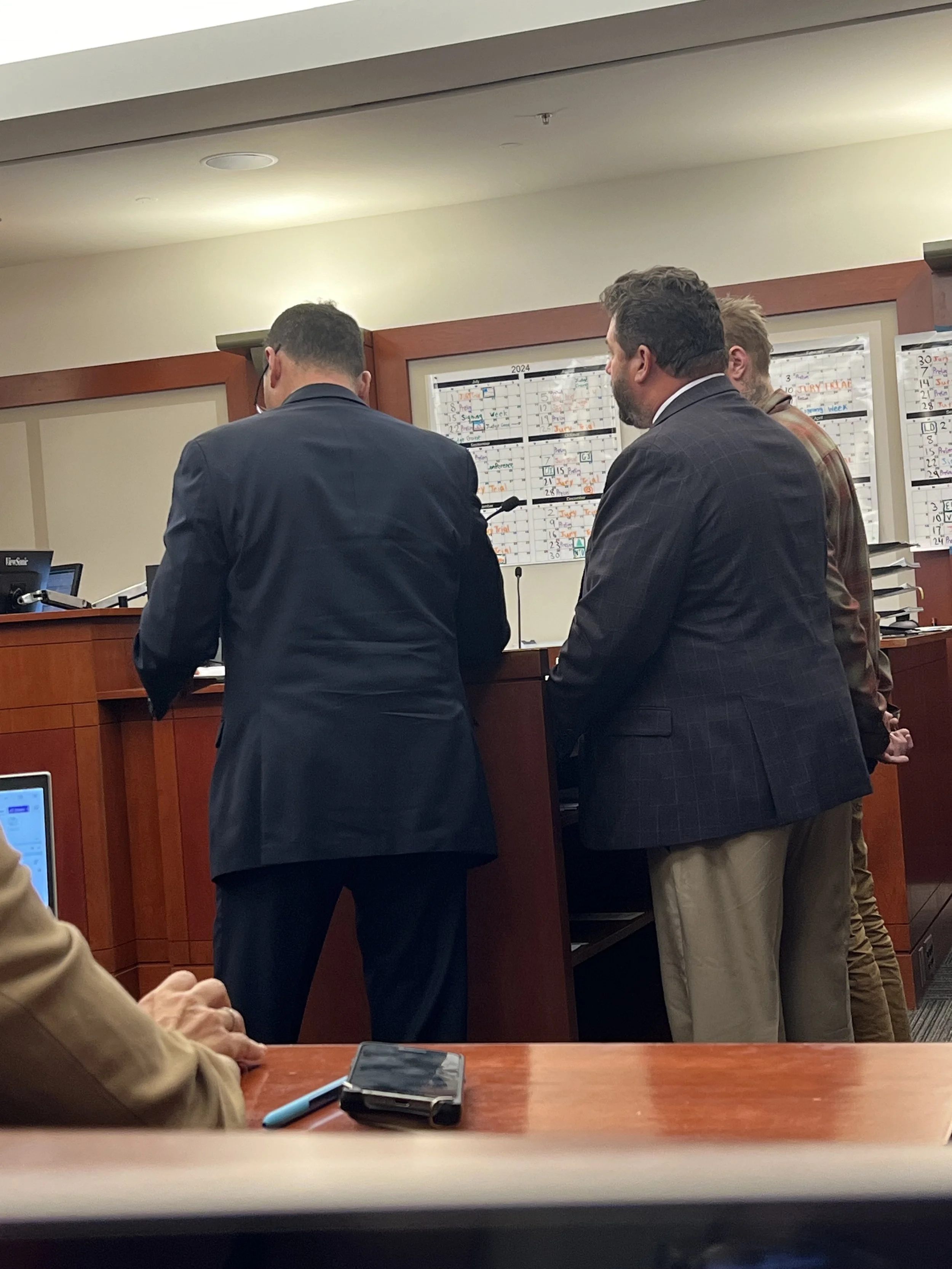 Several men in formal attire standing at a court counter with a large calendar on the wall behind them.